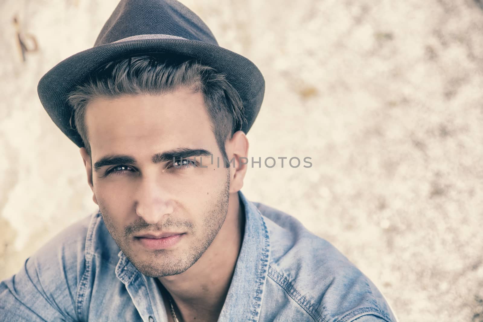 Handsome young, man in denim shirt and fedora hat sitting in pose on sandy beach and looking at camera