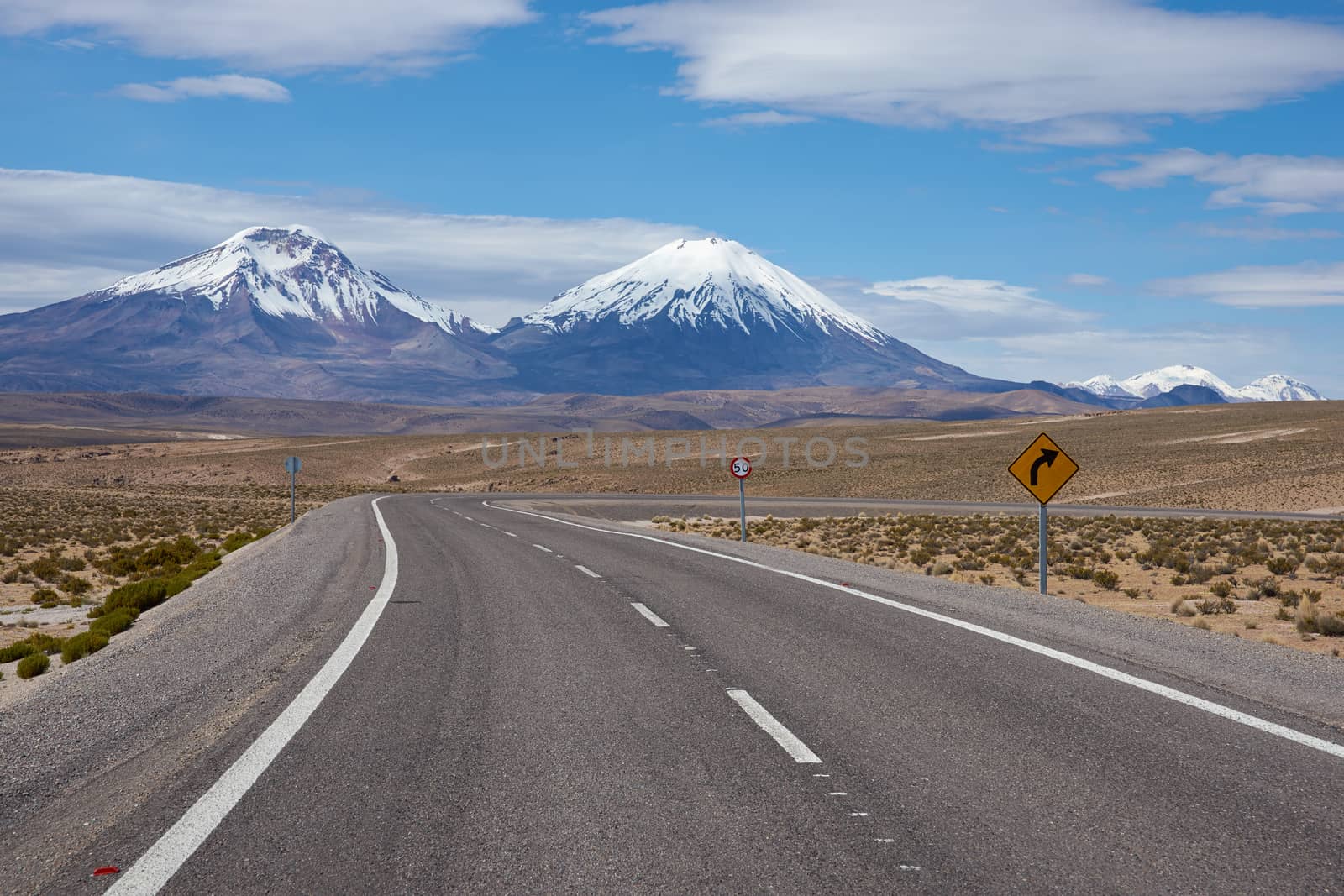 Road running towards the snow and ice covered peaks of the volcanoes Parinacota (6342m) and Pomerape (6240m) rising above the Altiplano of Northern Chile in Lauca National Park.