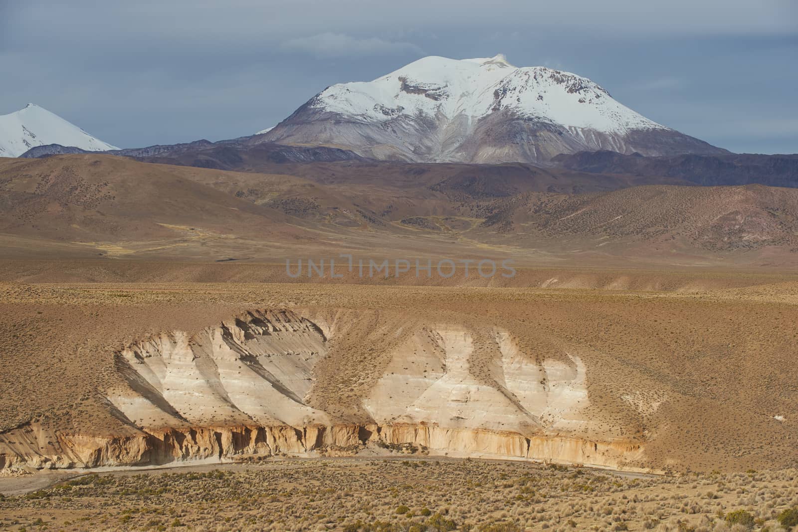 Snow covered peak of the active Guallatiri volcano (6063 m) towering above the Altiplano and cliffs running along the valley of the River Lauca in Lauca National Park, northern Chile.