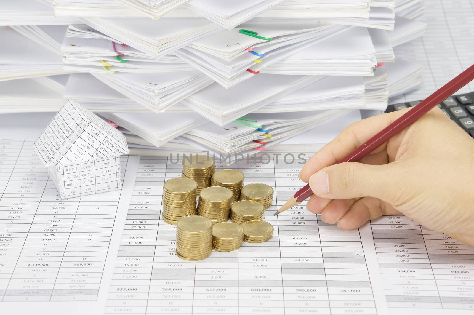 Man is auditing account by pencil with step pile of gold coins on the statement finance account have house with calculator and overload of document with colorful paperclip as background.