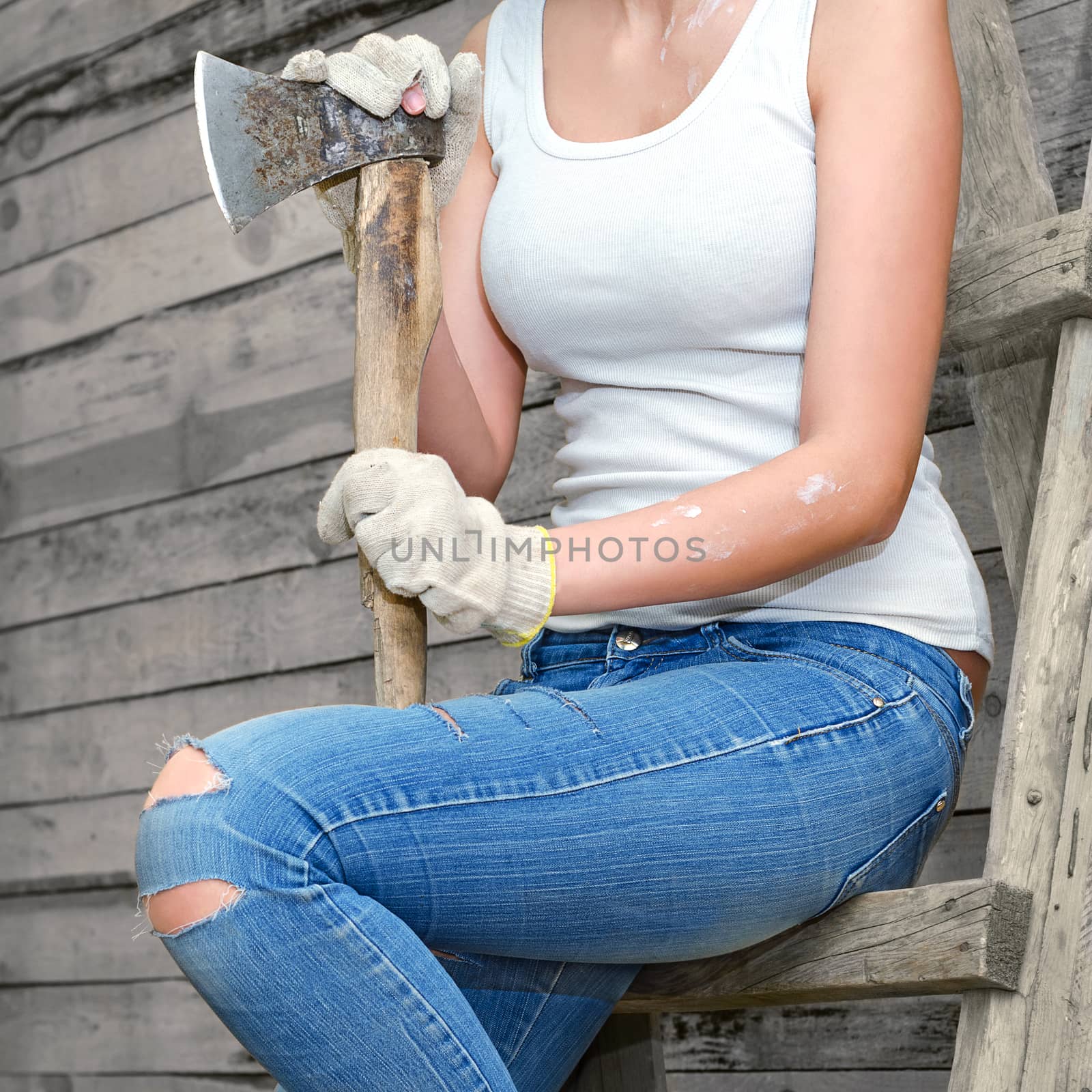 Girl sitting on an old ladder against the wall from the old boards. In old clothes and work gloves, holding an old axe.