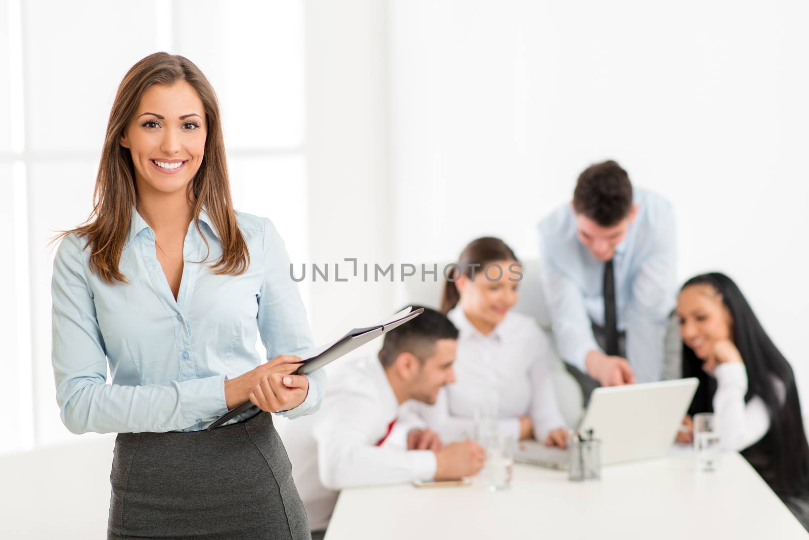 Successful young businesswoman standing proudly in the office and looking at the camera. Her young colleagues working at laptop on desk.