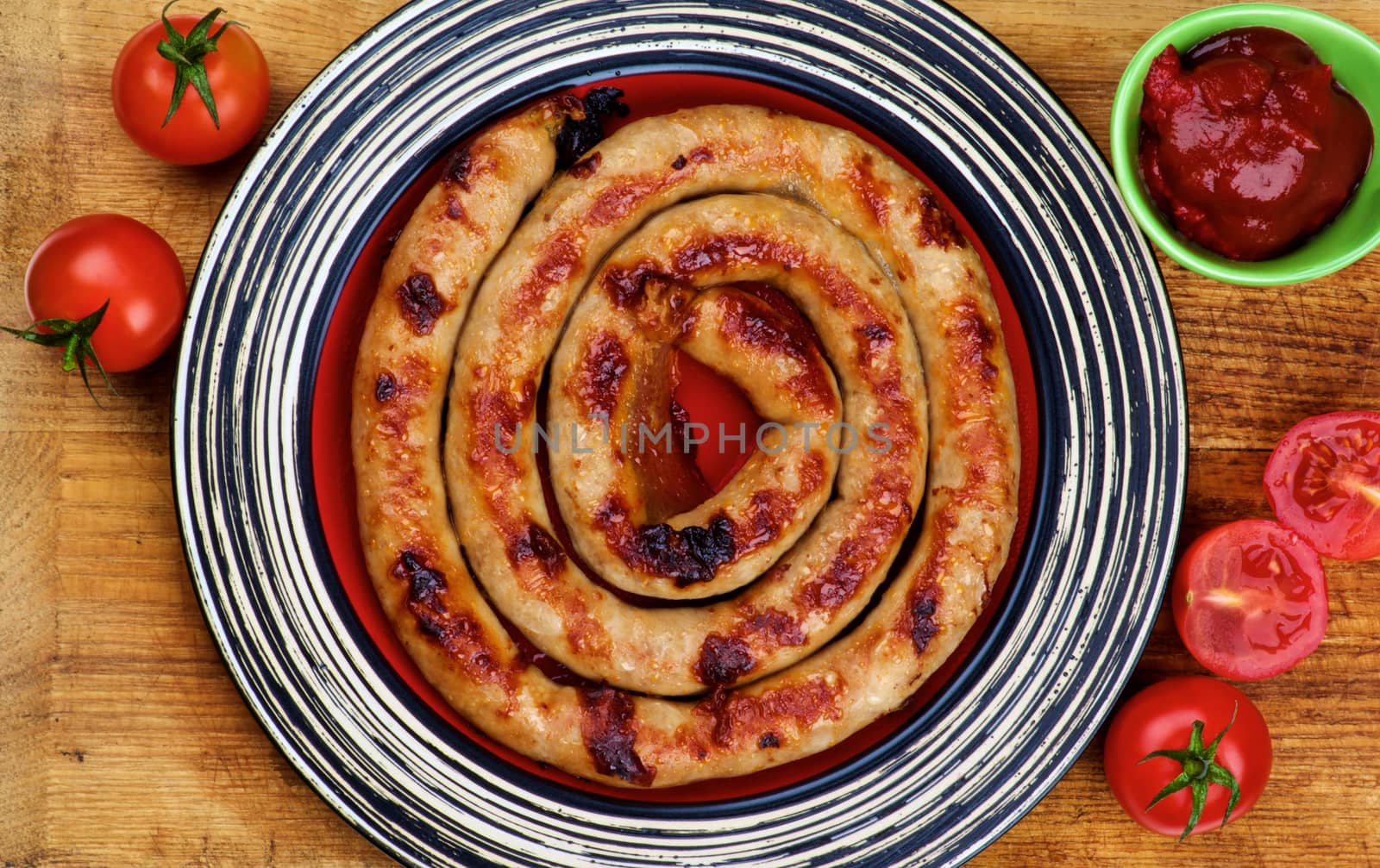 Delicious Grilled Spiral Sausage on Striped Plate with Ketchup and Fresh Cherry Tomatoes closeup on Wooden Cutting Board. Top View 