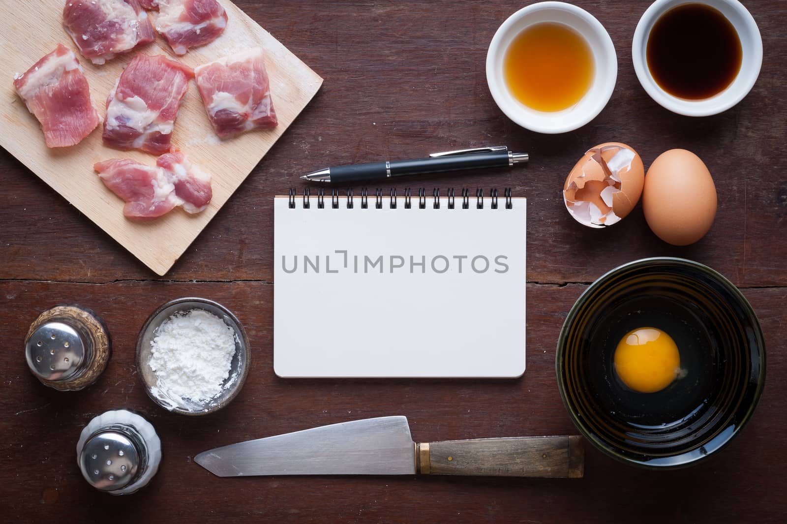 Preparation for cooking in home kitchen. Soft pork ribs on chopping board, notepad for check list, egg, and ingredients on wood table.