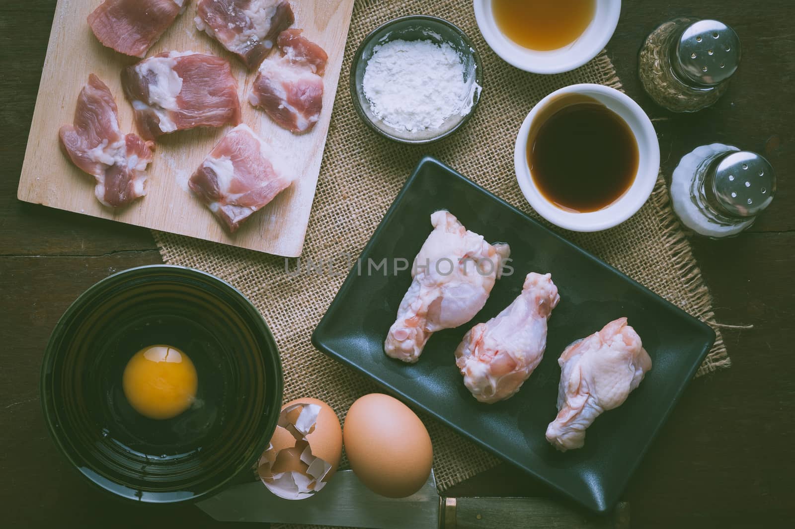 Preparation for cooking in home kitchen. Soft pork ribs on chopping board, chicken wings in ceramic dish, eggs, and ingredients on wood table with vintage filter effect.