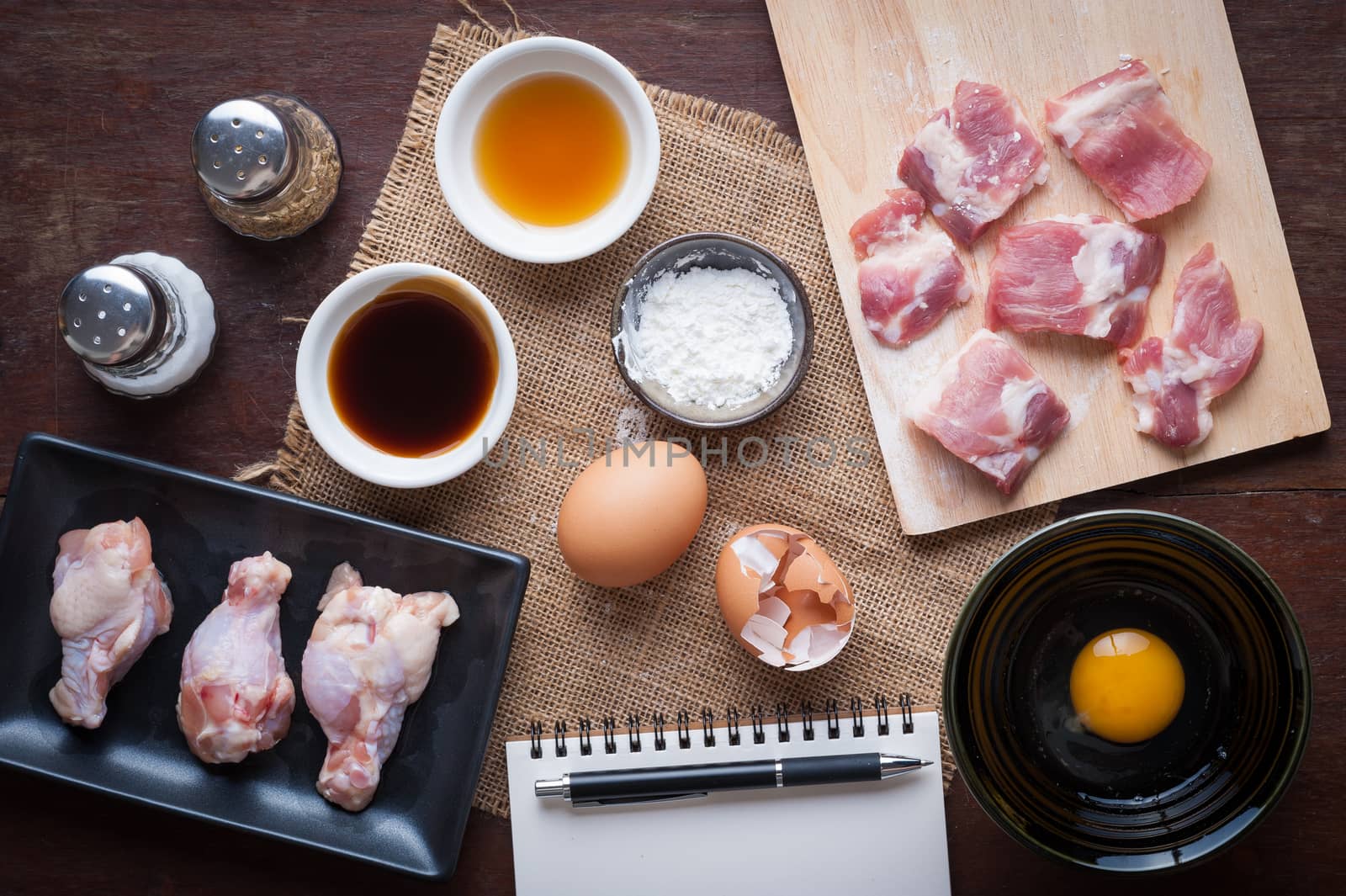 Preparation for cooking in home kitchen. Soft pork ribs on chopping board, chicken wings in ceramic dish, eggs, and ingredients on wood table.
