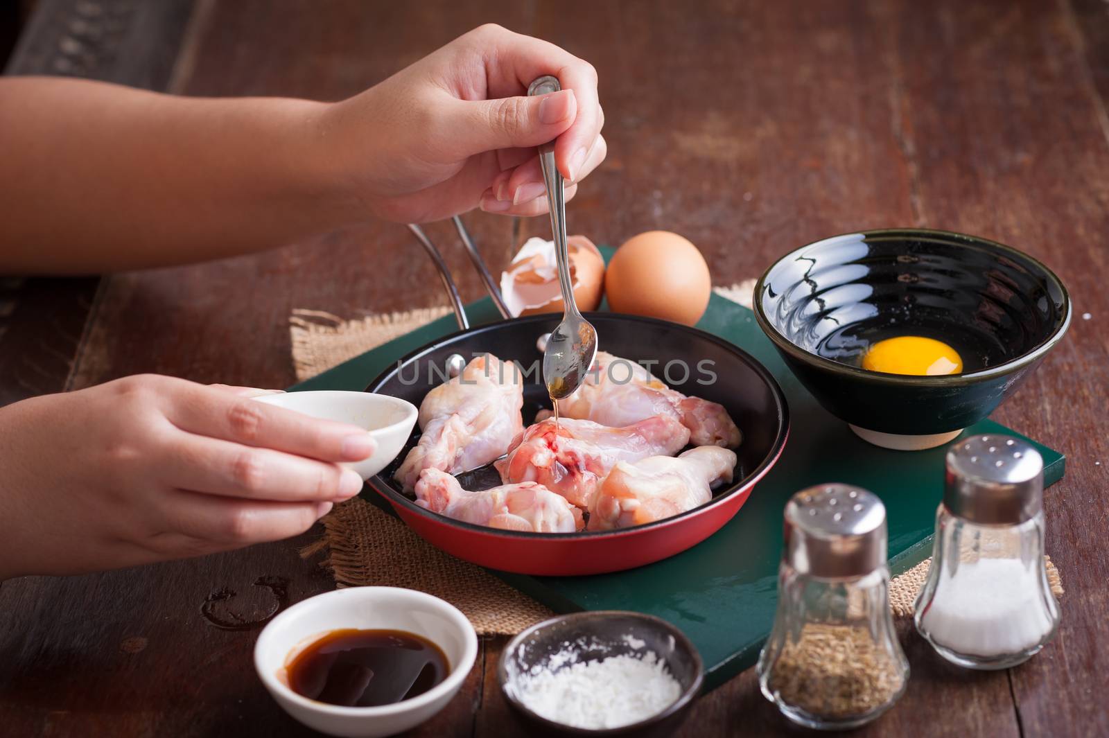 Preparation for cooking in home kitchen. Woman left hand pouring oyster sauce into fry pan to mix with chicken wings.
