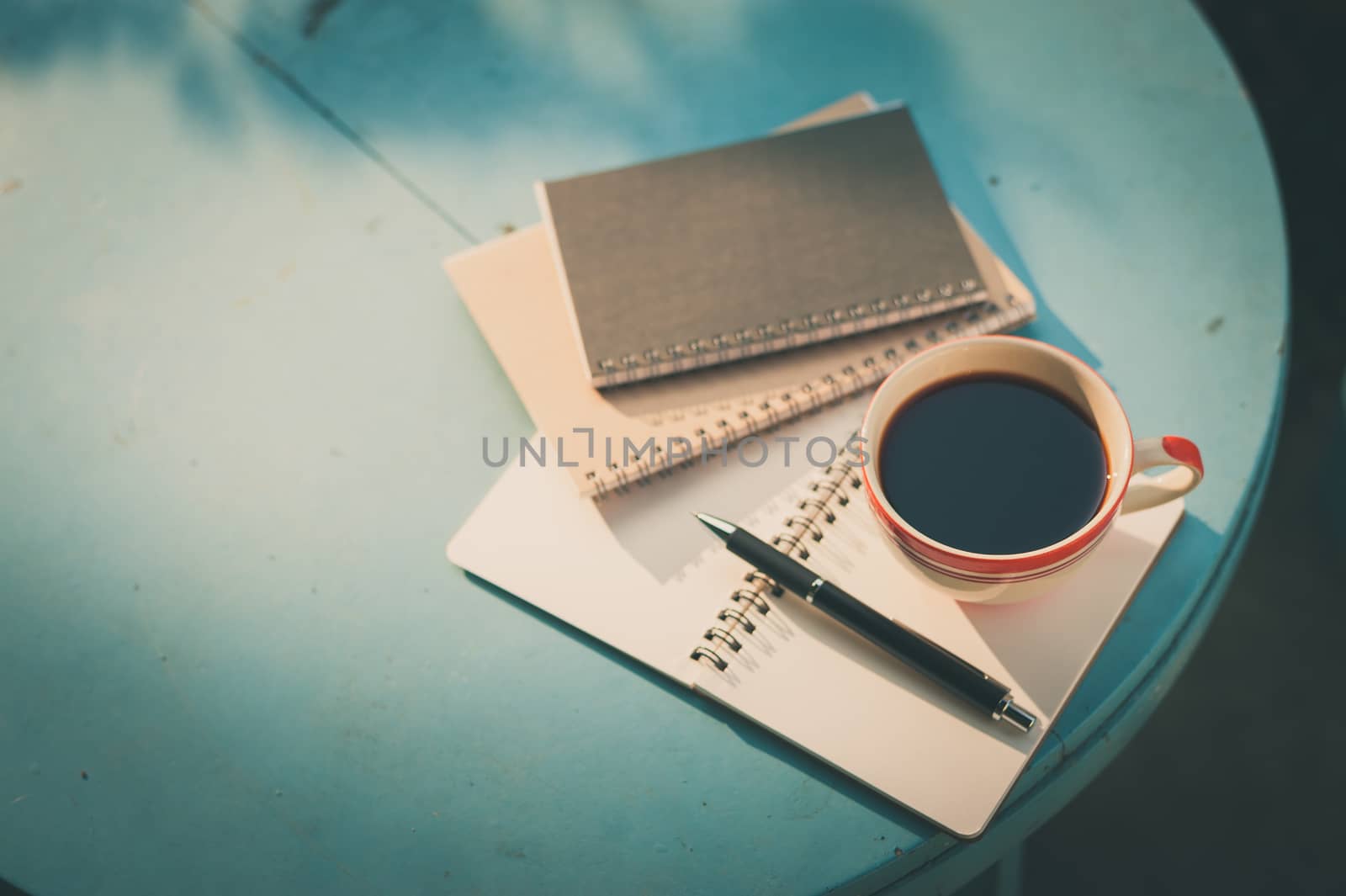 Coffee cup with notebooks and pen on blue round wood table in morning time with film filter effect