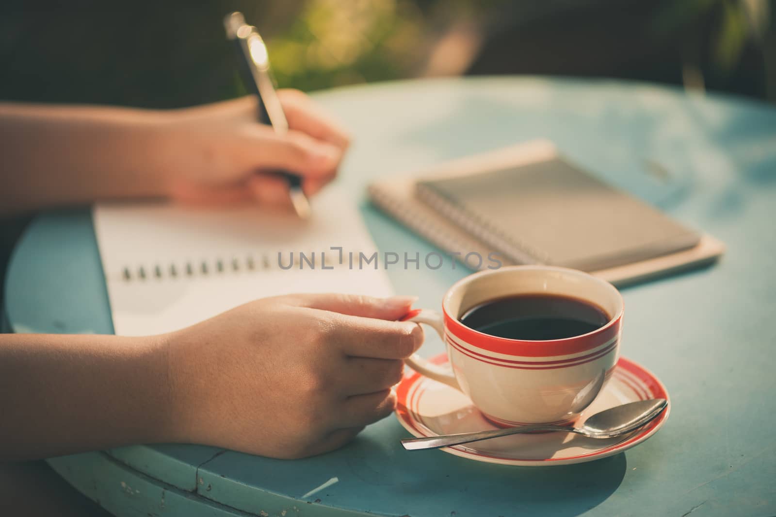 Woman right hand holding coffee cup while writing on notebook at outdoor area in cafe with vintage filter effect