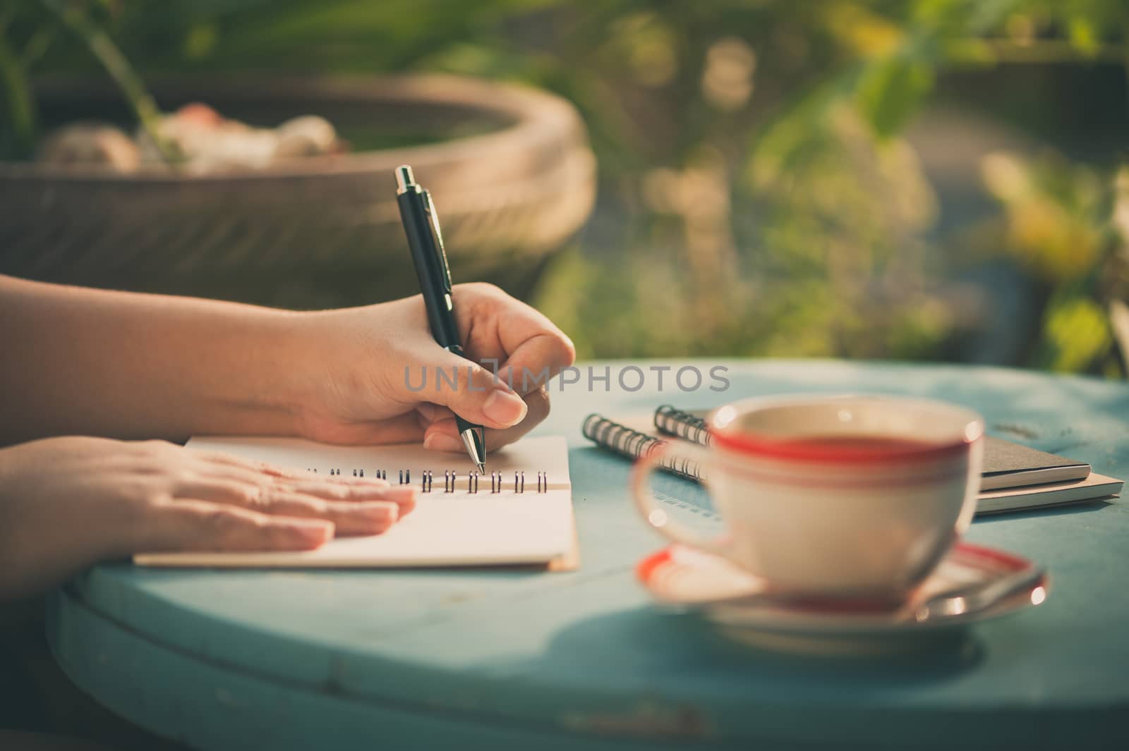 Woman left hand writing journal on small notebook in outdoor area at cafe with morning scene and vintage filter effect