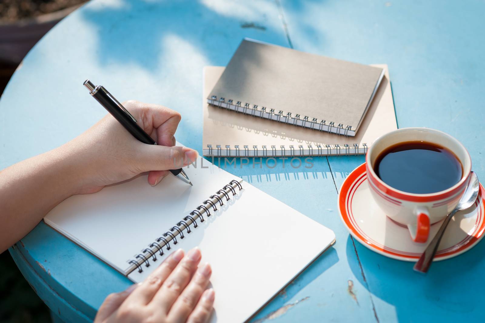 Woman left hand writing journal on small notebook in outdoor area at cafe with morning scene