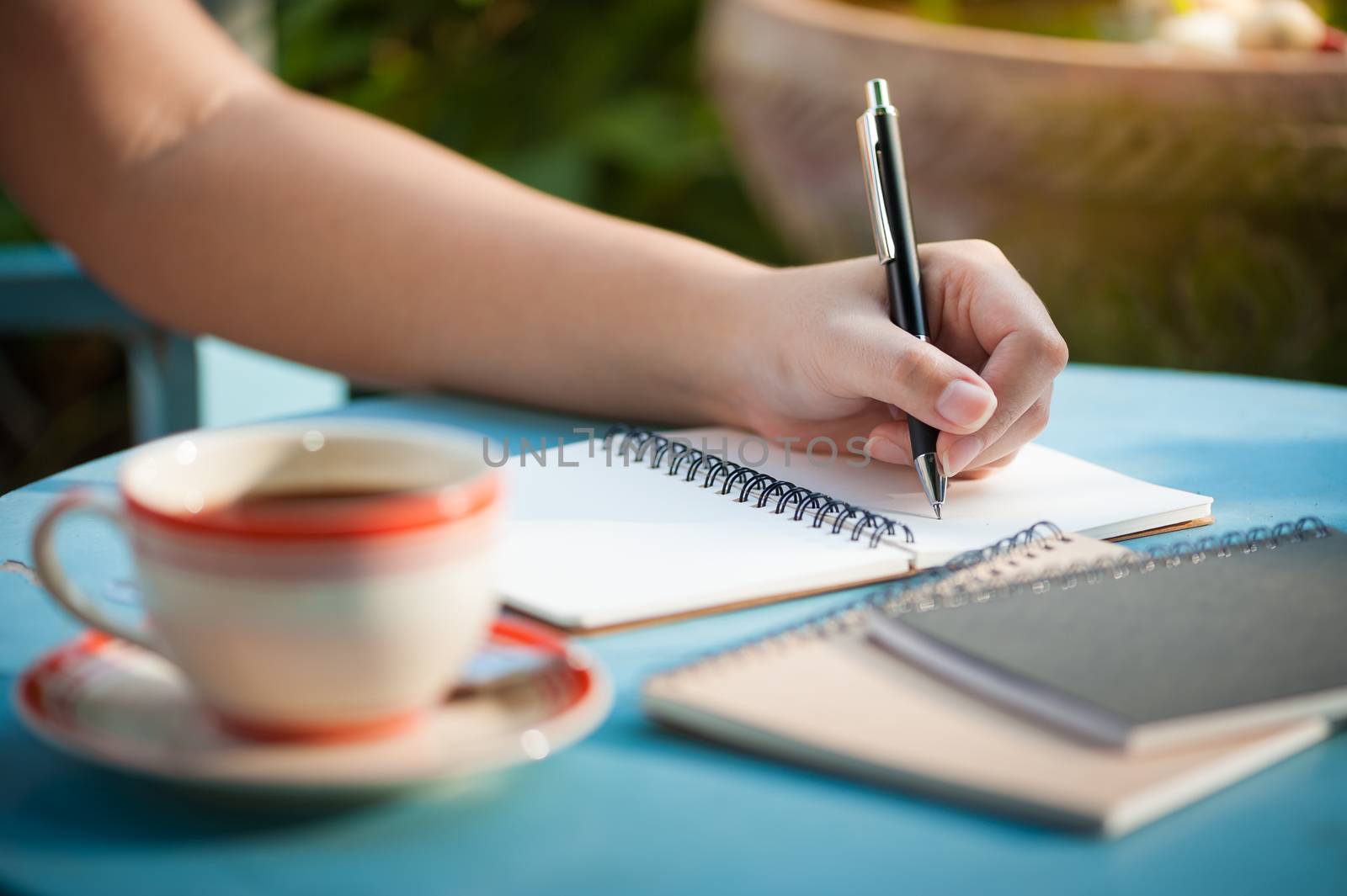 Woman left hand writing journal on small notebook in outdoor area at cafe with morning scene