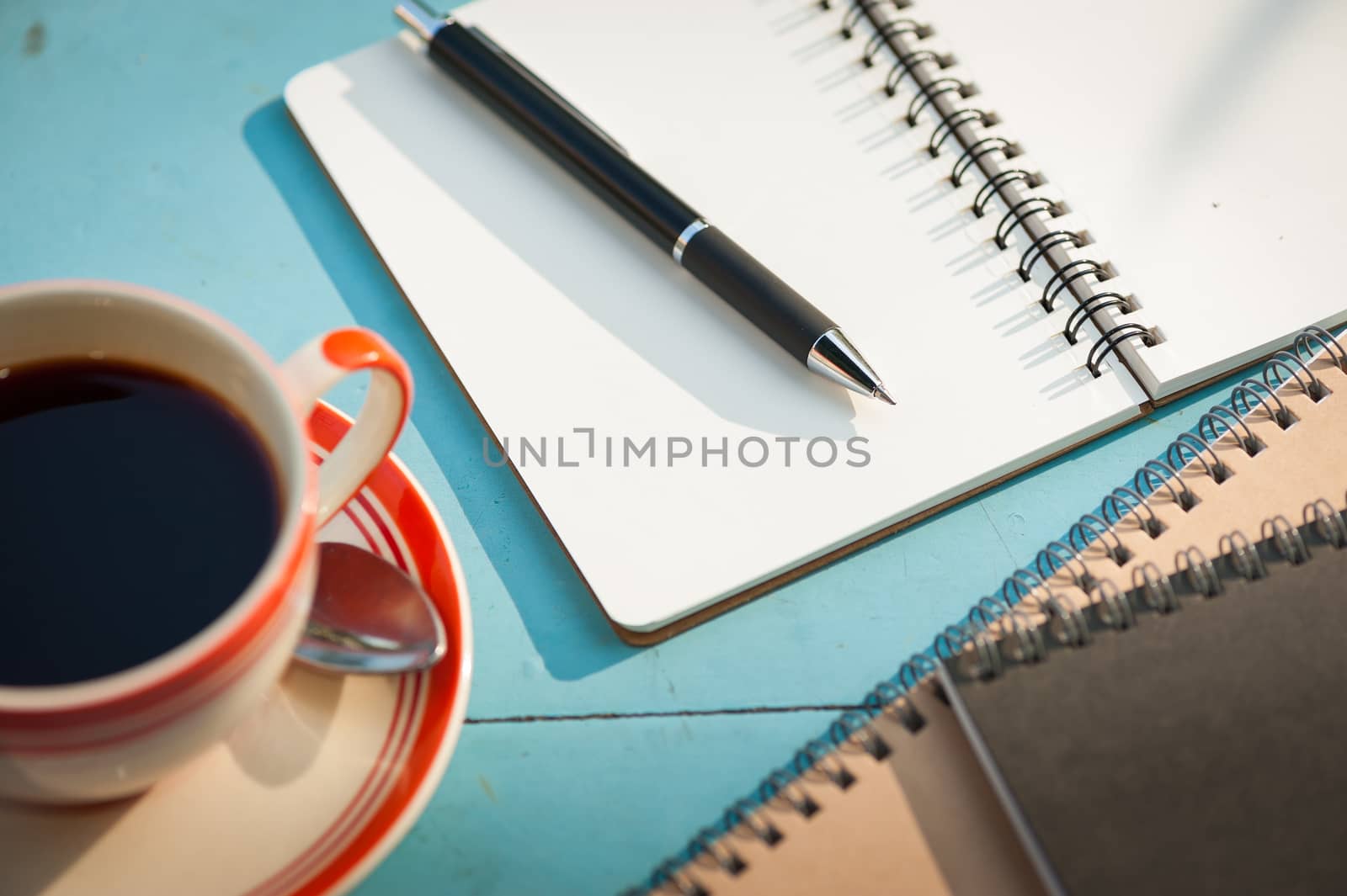 Small notebook and pen beside coffee cup on blue wood table at outdoor area in cafe with morning scene