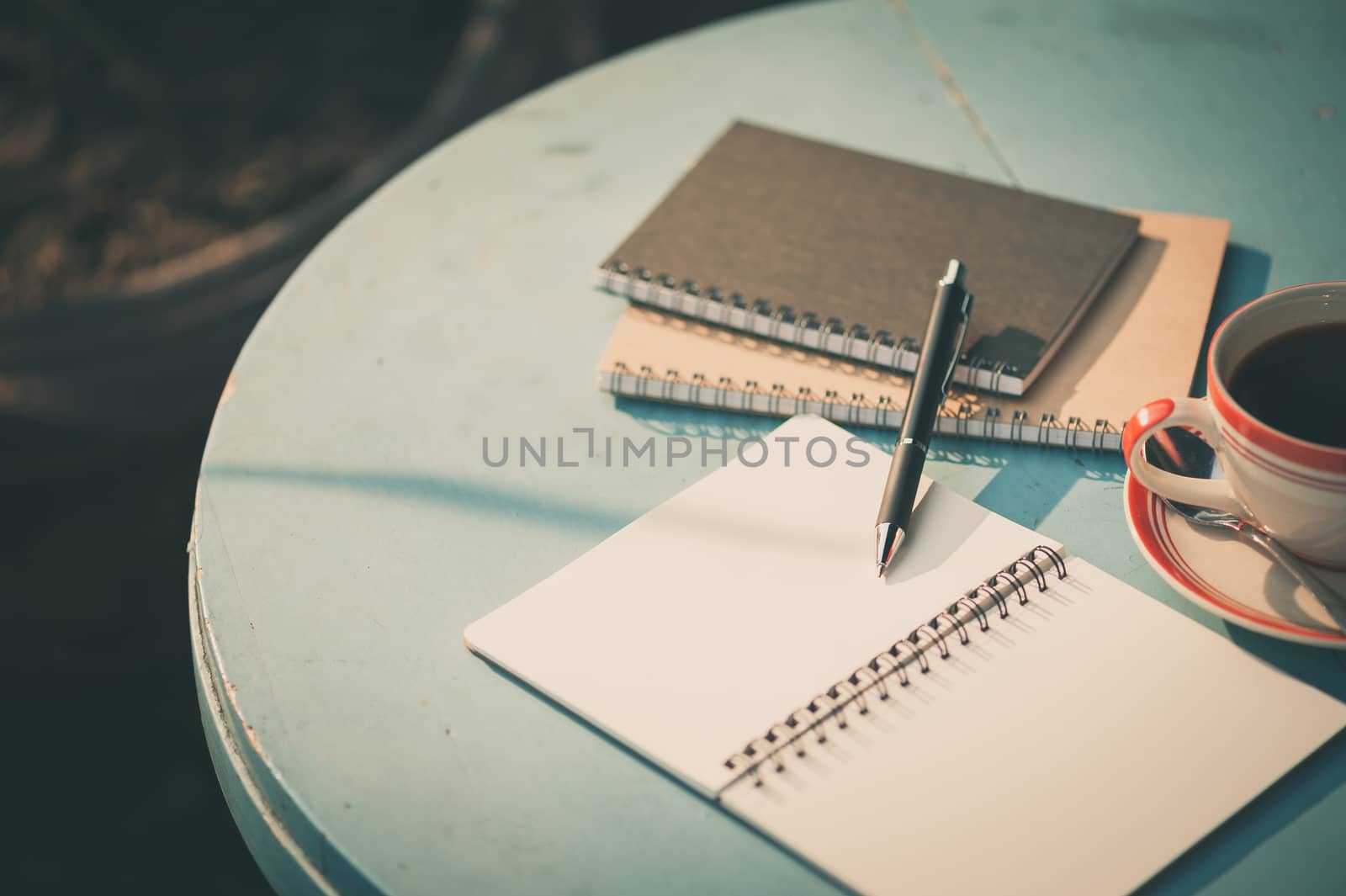 Small notebook and pen beside coffee cup on blue color round wood table at outdoor area in cafe with morning scene and vintage filter effect