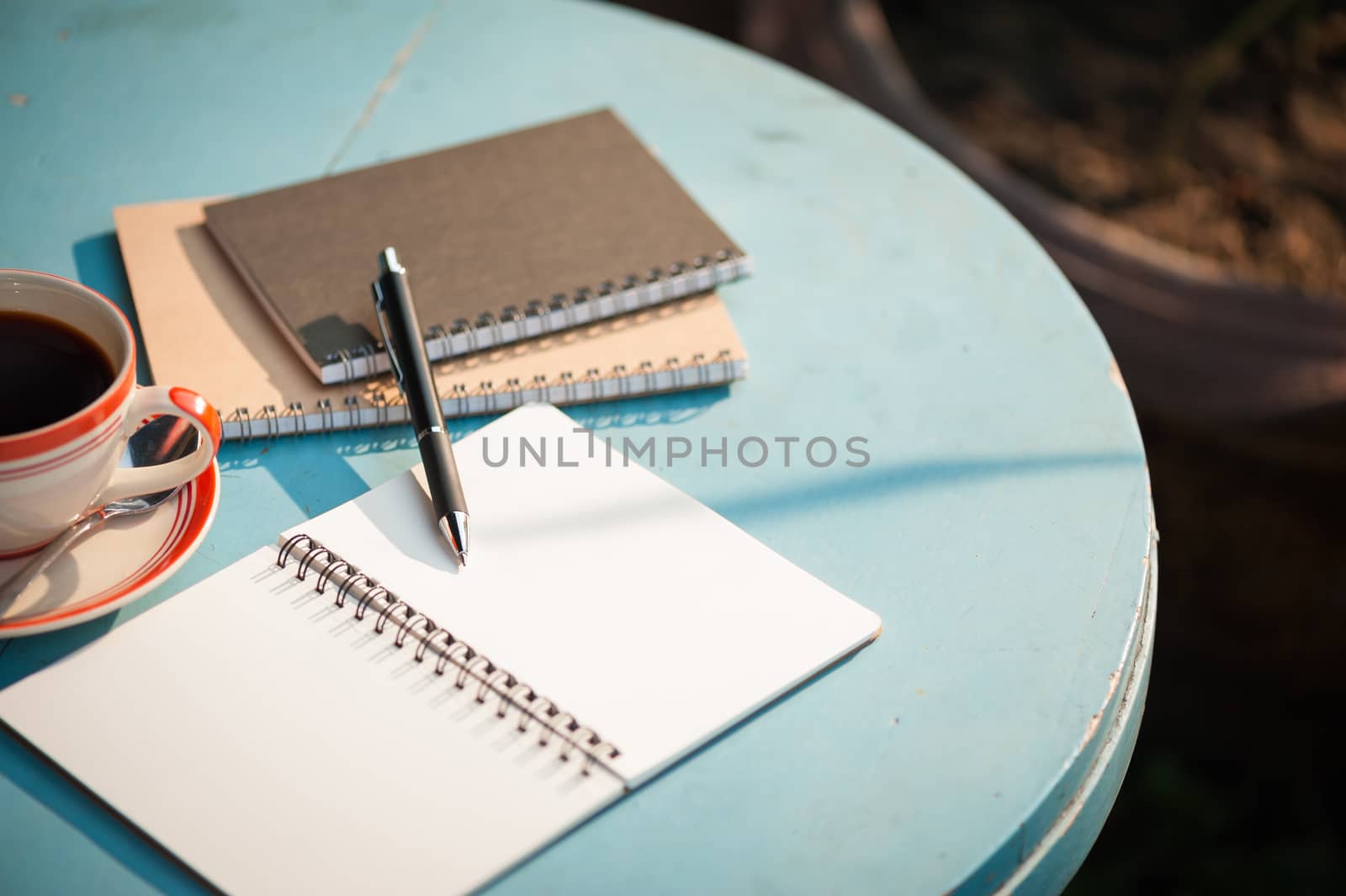 Small notebook and pen beside coffee cup on blue color round wood table at outdoor area in cafe with morning scene