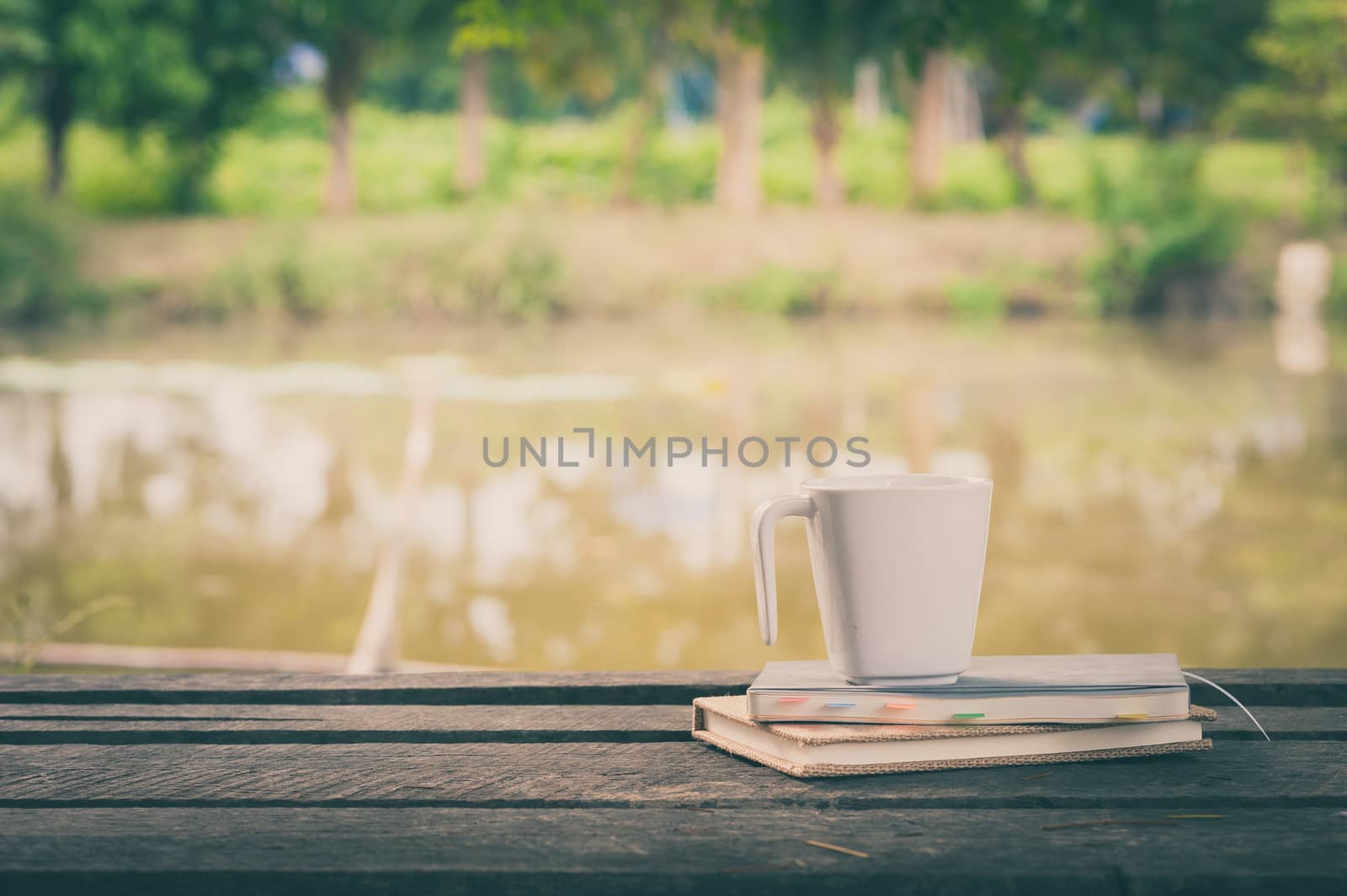 Coffee cup and notebooks on rustic wood table at outdoor area with rural lake view on blurry background in morning time with vintage filter effect