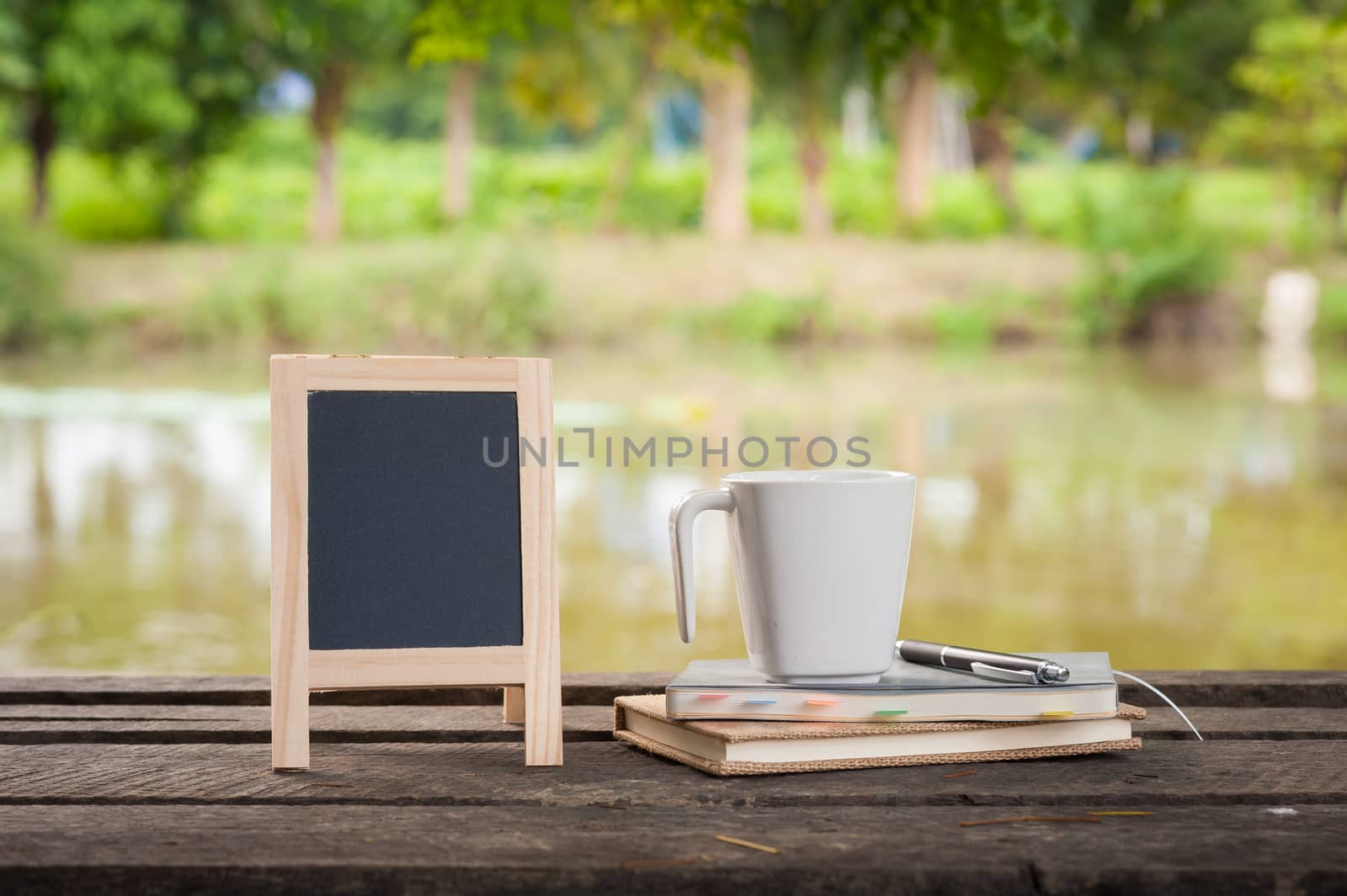 Small A-frame blackboard with blank area for text or message, notebooks and coffee cup on rustic wood table with rural lake view in blurry background at morning time