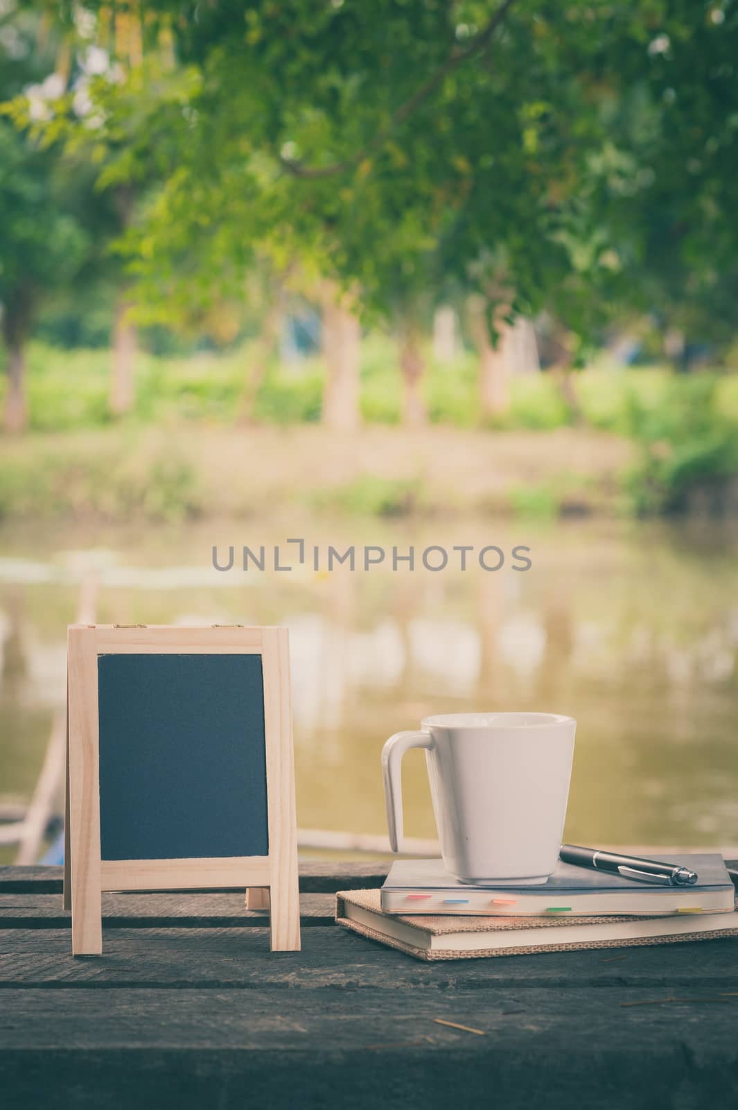 Small A-frame blackboard with blank area for text or message, notebooks and coffee cup on rustic wood table with rural lake view in blurry background at morning time with vintage filter effect