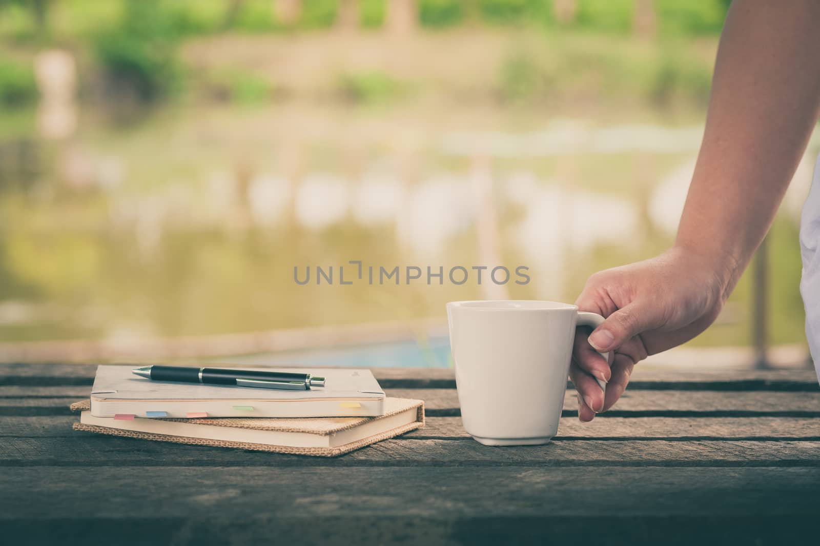 Woman hand holding coffee cup beside notebooks on rustic wood bench with rural lake view in blurry background on weekend with morning scene and vintage filter effect