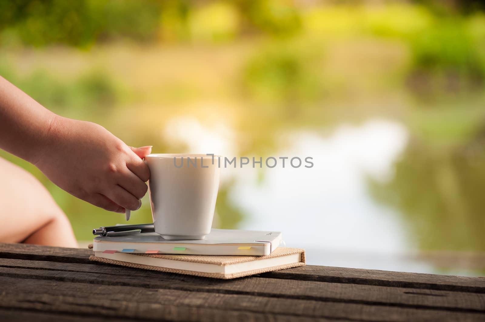 Woman right hand holding coffee cup beside notebooks on rustic wood bench in morning time on weekend