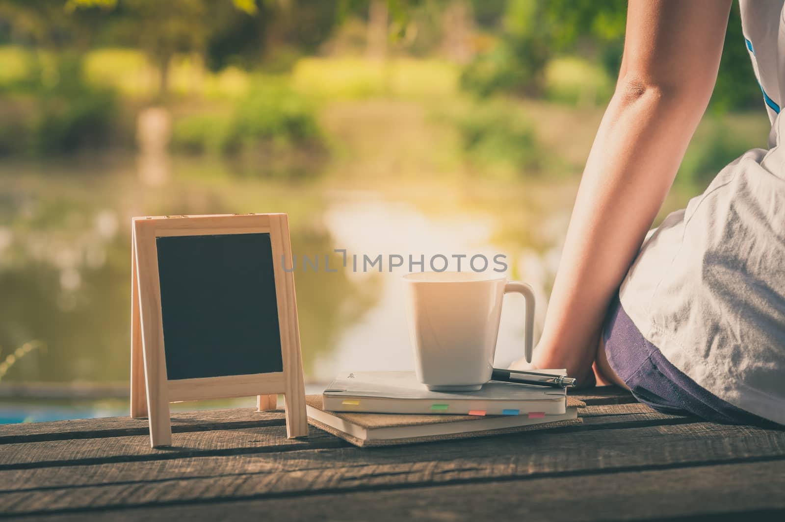 Young woman sitting and put down her hand beside coffee cup, notebooks and small A-frame blackboard on rustic wood bench in weekend with morning scene and vintage filter effect