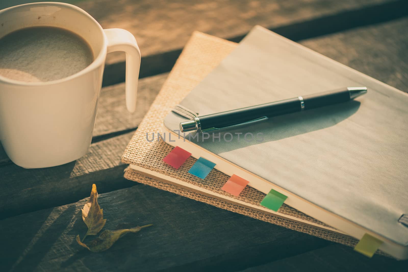 Pen, notebooks, and coffee cup on rustic wood table at outdoor area in morning time on summer with vintage filter effect