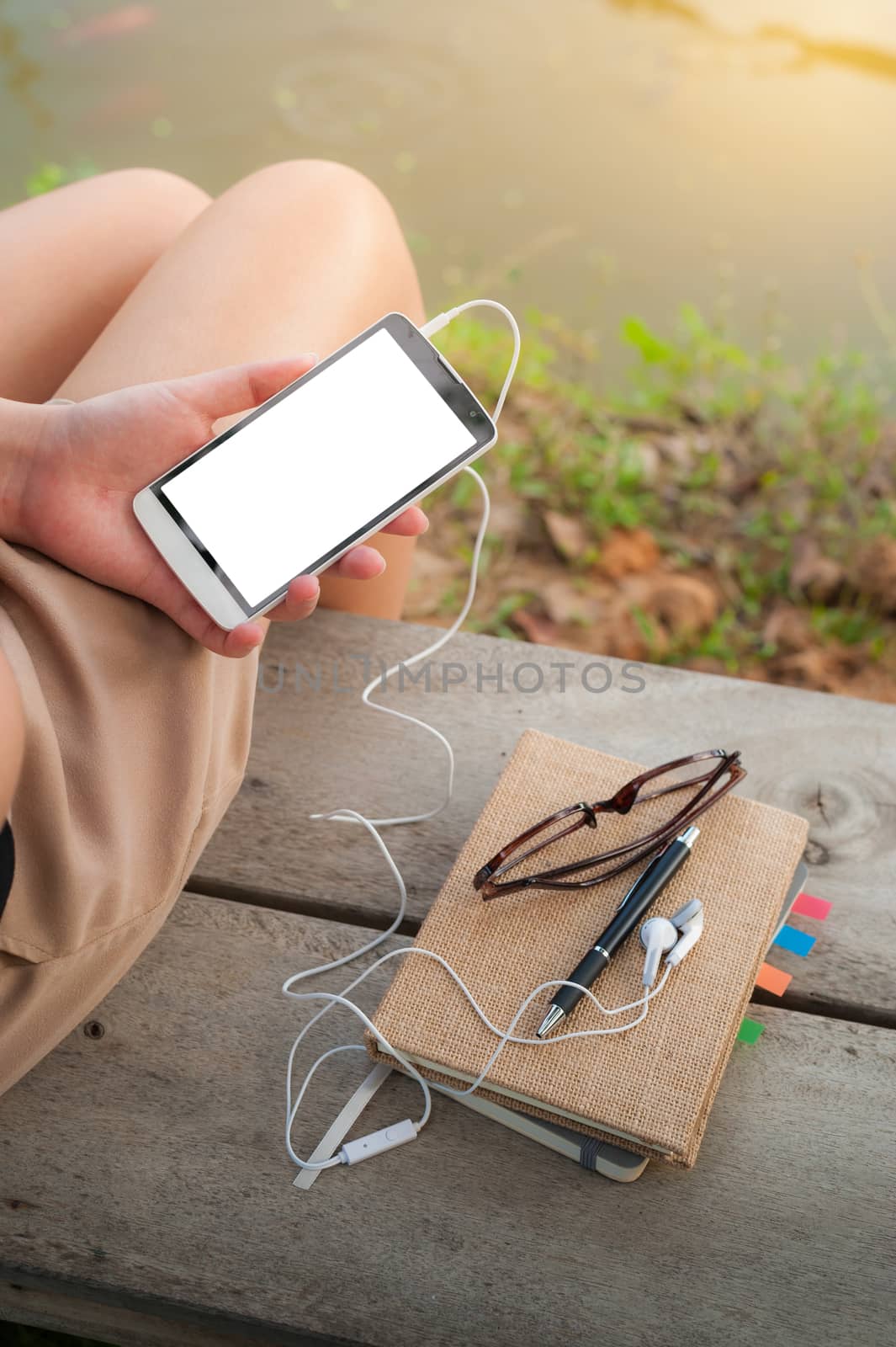 Young woman holding smartphone with small headphones by left hand while sitting on wood bench in park in morning time on weekend