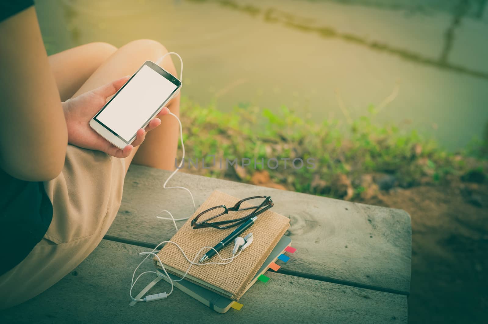 Young woman holding smartphone with small headphones by left hand while sitting on wood bench in park in morning time on weekend with vintage filter effect