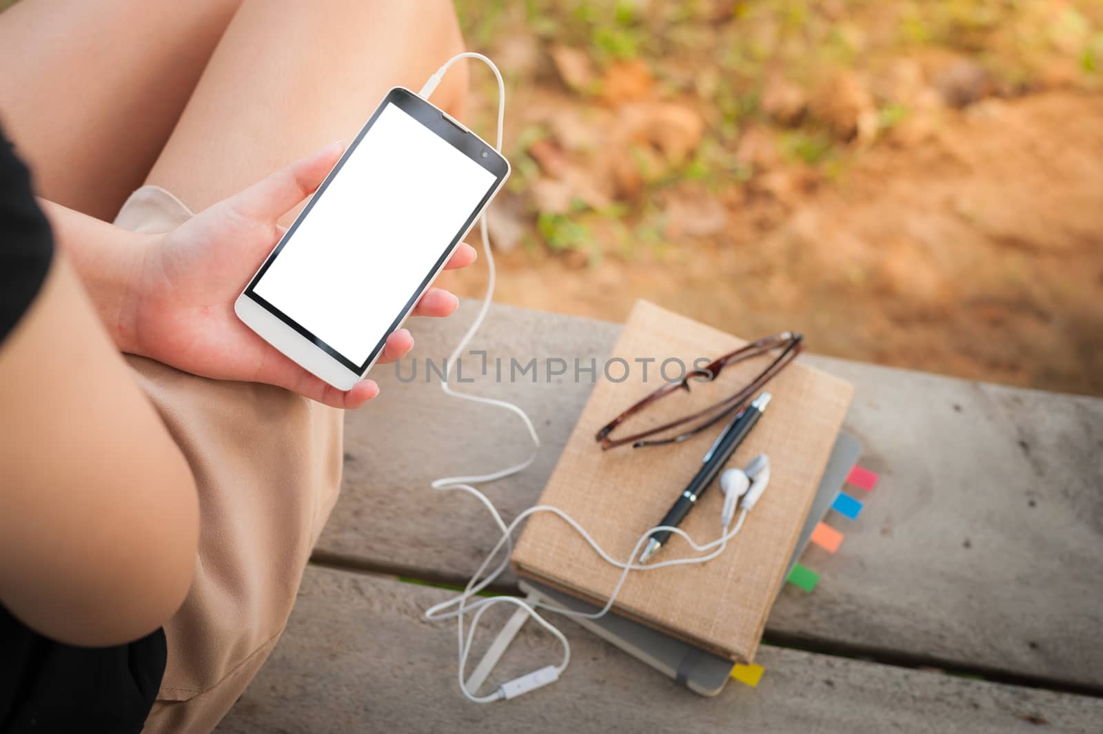 Young woman holding smartphone with small headphones by left hand while sitting on wood bench in park in morning time on weekend
