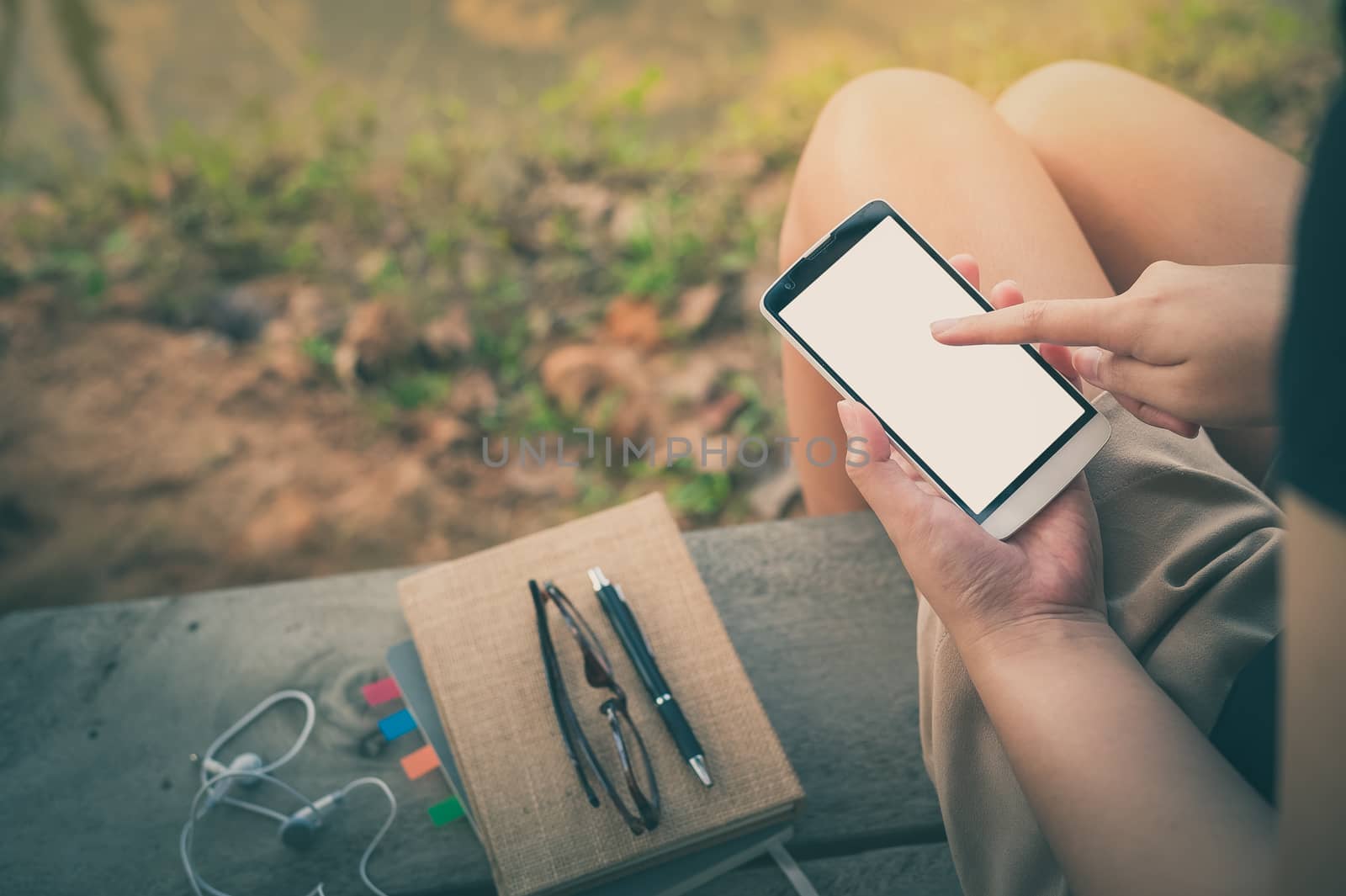 Young woman touching on smartphone blank screen while sitting on wood bench in park in morning time on weekend with vintage filter effect