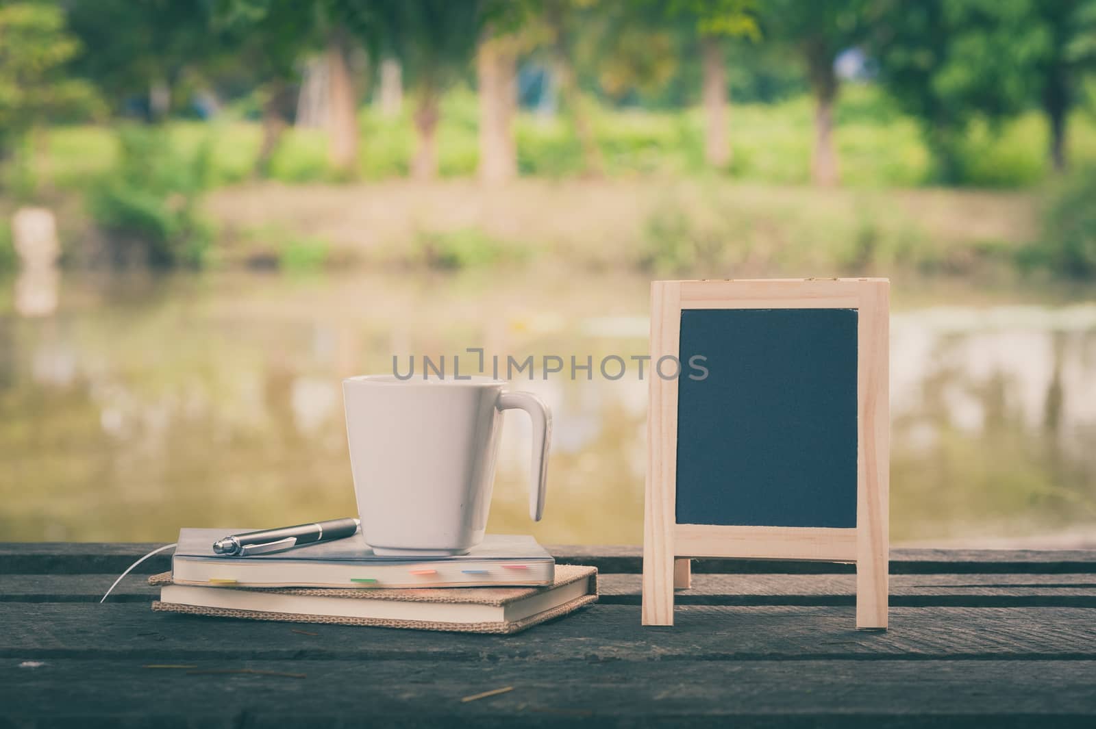 Small A-frame blackboard with blank area for text or message, notebooks and coffee cup on rustic wood table with rural lake view in blurry background at morning time with vintage filter effect