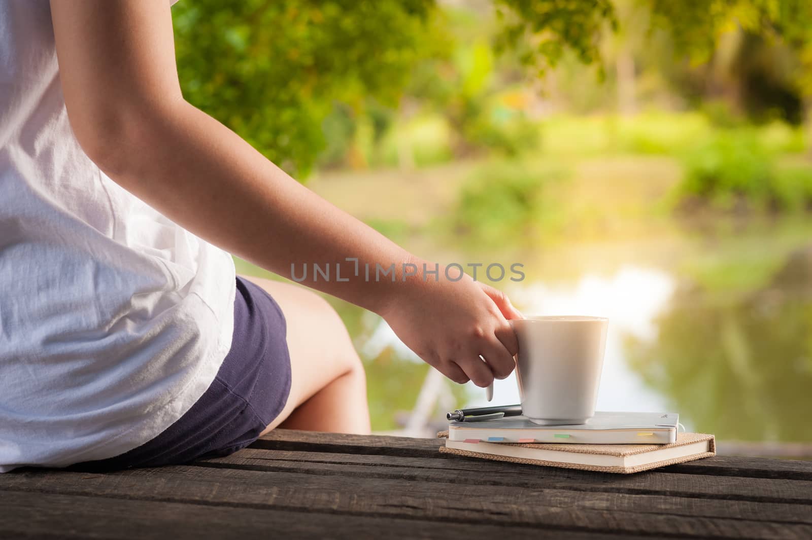 Woman right hand holding coffee cup beside notebooks on rustic wood bench with rural lake view in background in morning time on weekend