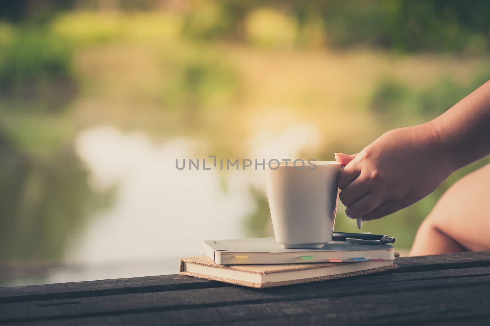 Woman left hand holding coffee cup beside notebooks on rustic wood bench in morning time on weekend with vintage filter effect