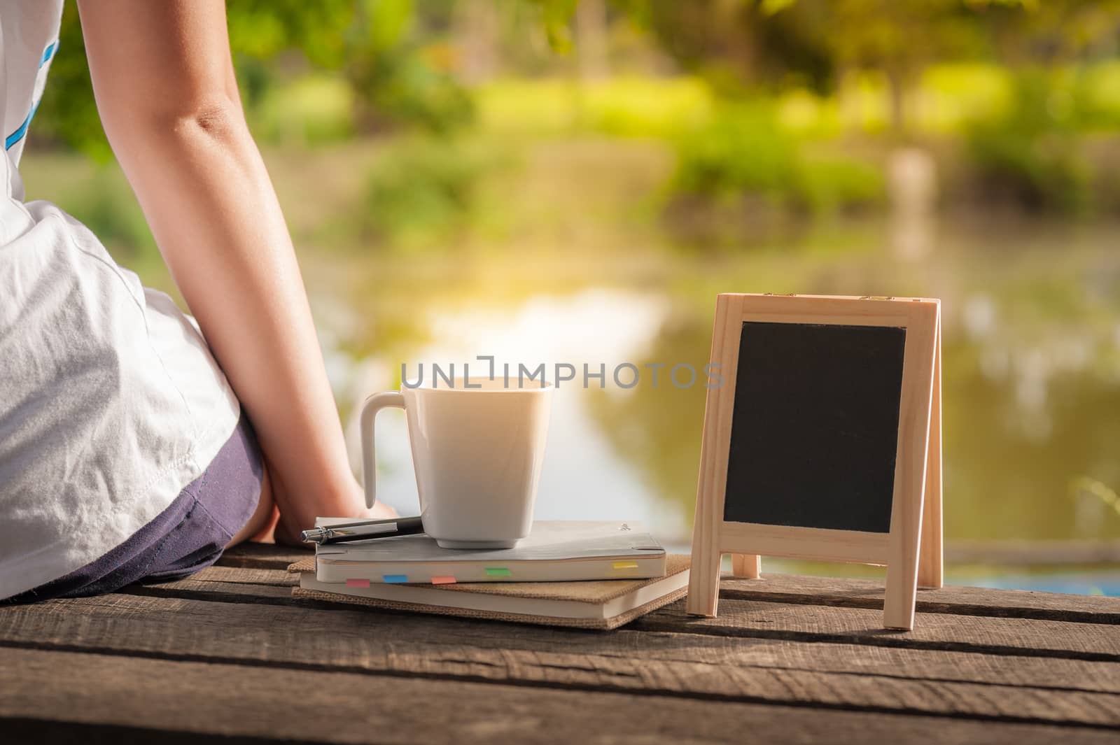 Young woman sitting and put down her hand beside coffee cup, notebooks and small A-frame blackboard on rustic wood bench in weekend with morning scene