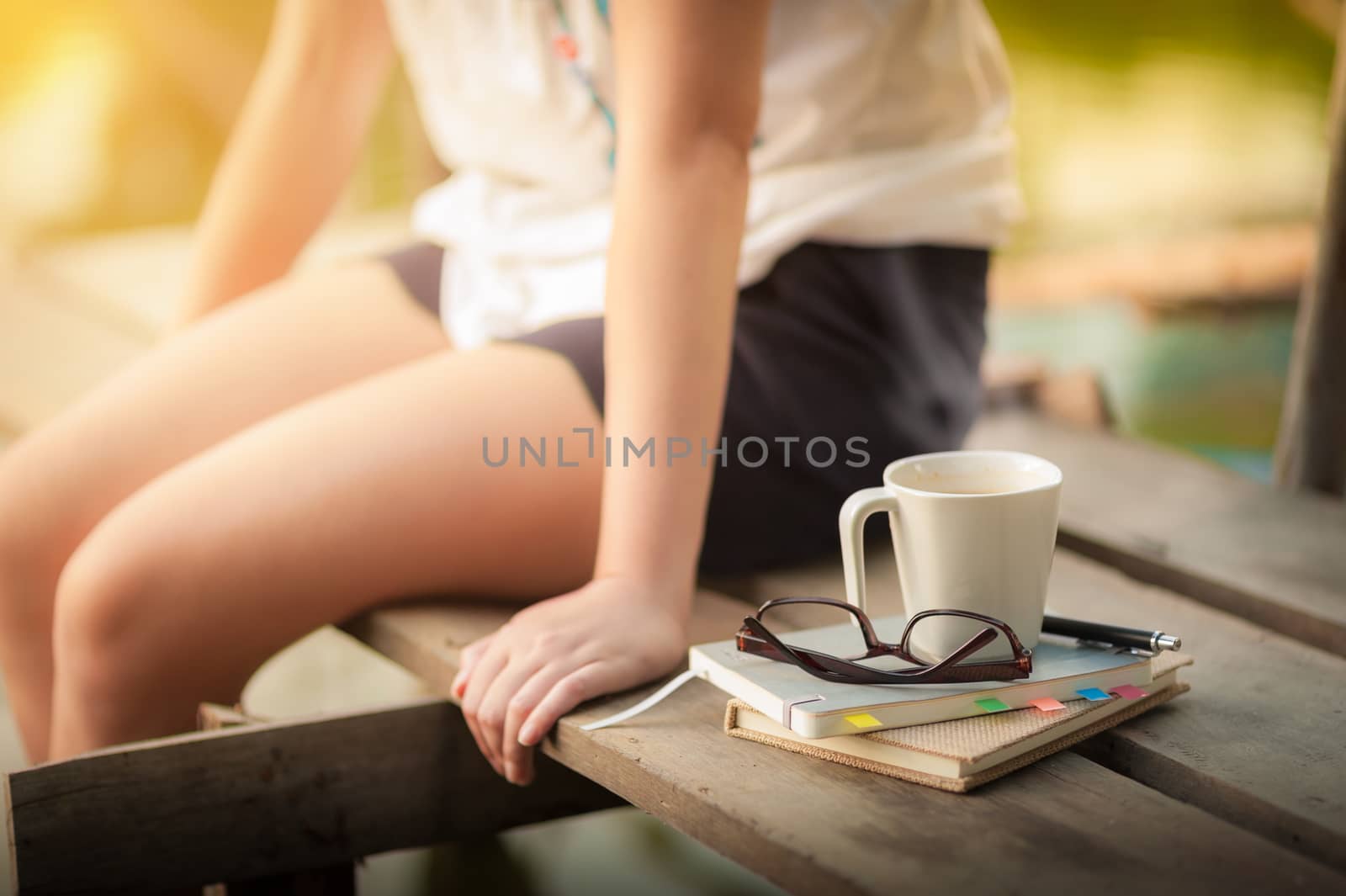 Notebooks, pen, glasses, and coffee cup are putting down on the wood waterside while young woman sitting beside them in weekend with morning scene