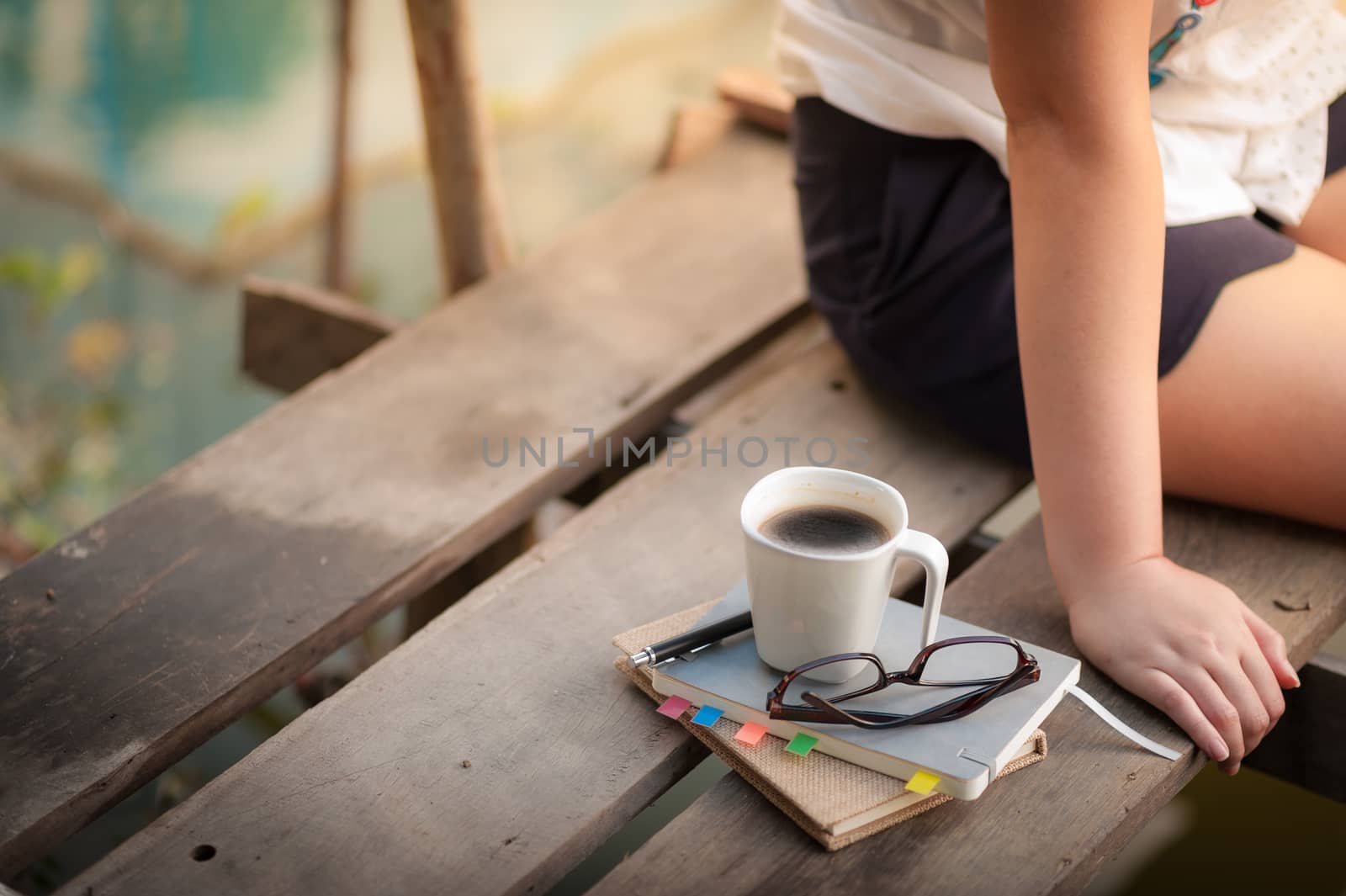 Notebooks, pen, glasses, and coffee cup are putting down on the wood waterside while young woman sitting beside them in morning time on weekend