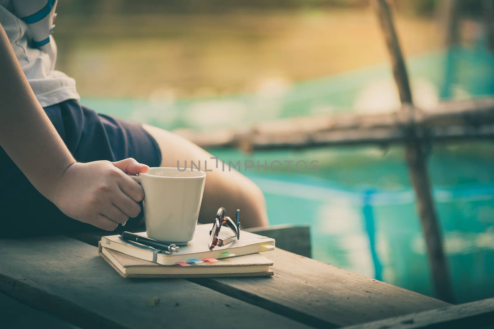 Young woman right hand holding coffee cup above notebokks on wood waterside in morning time on weekend