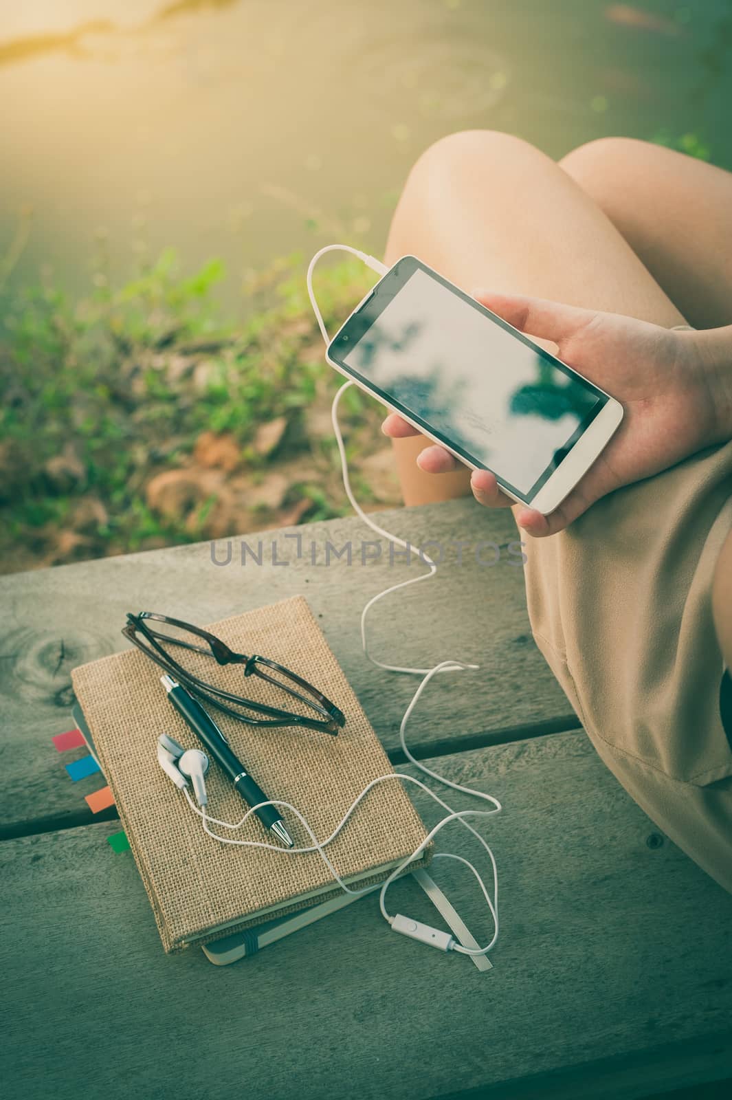 Young woman  holding smartphone with small headphones by right hand while sitting on wood bench in park in morning time on weekend with vintage filter effect