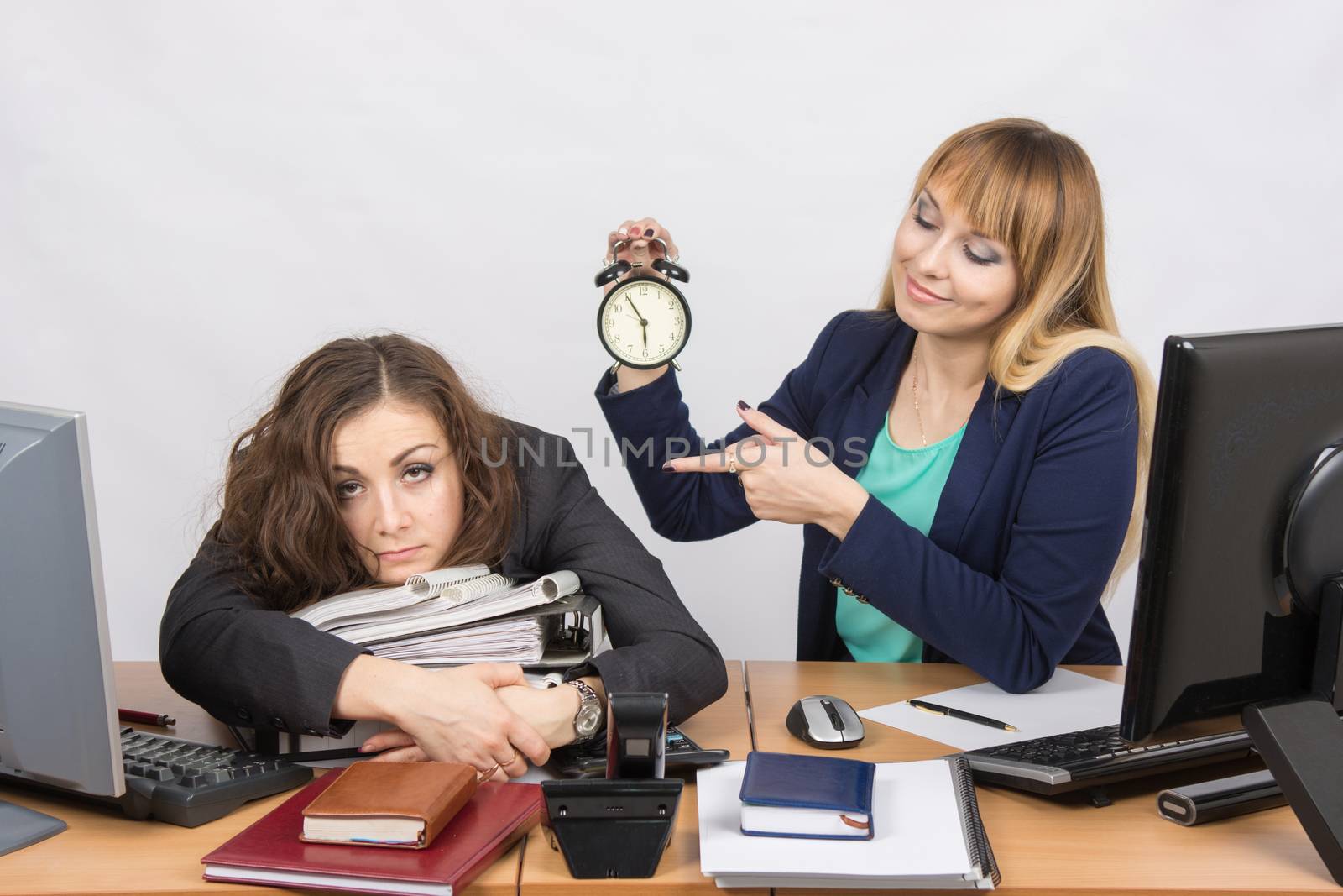 The girl in the office with a smile, holding a clock and looking at the tormented colleague