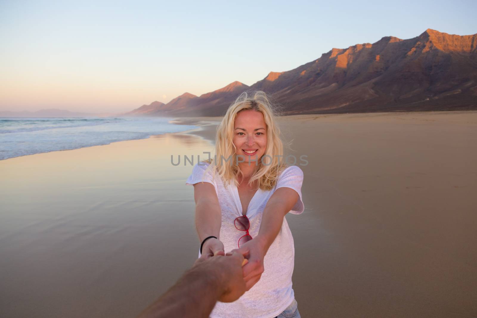 Young romantic couple, holding hands, having fun on perfect deserted beach at sunset. Shot from boyfrieds perspective. Guy looking at her beautiful carefree girlfriend.