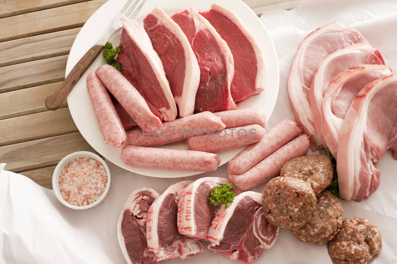 Assorted raw meat cuts with salt including pork cutlets, steak, lamb chops, sausage and meatballs on a wooden table