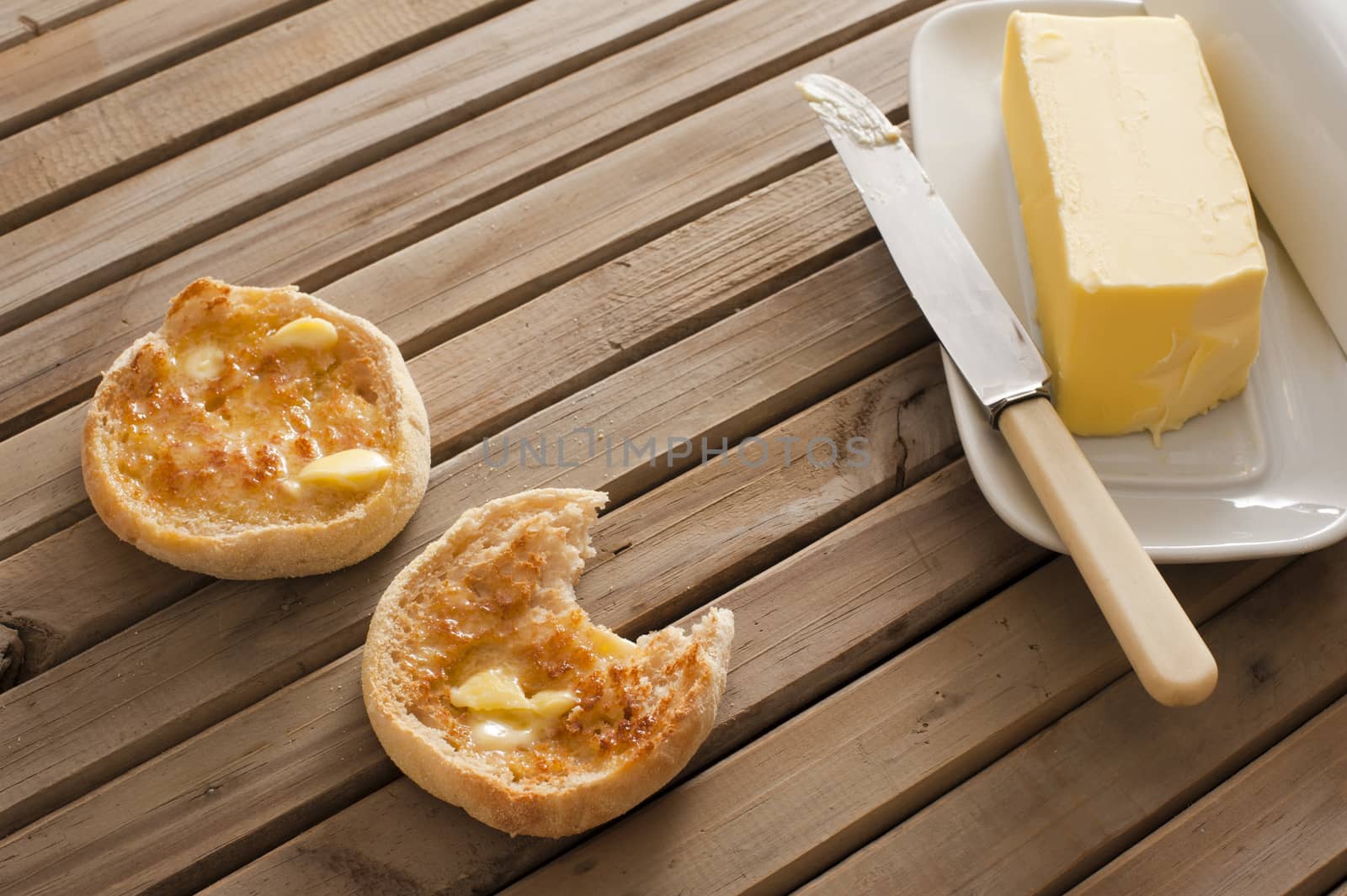 Buttered crumpets for breakfast with a pat of butter alongside on a plate served on a wooden table, one crumpet bitten into