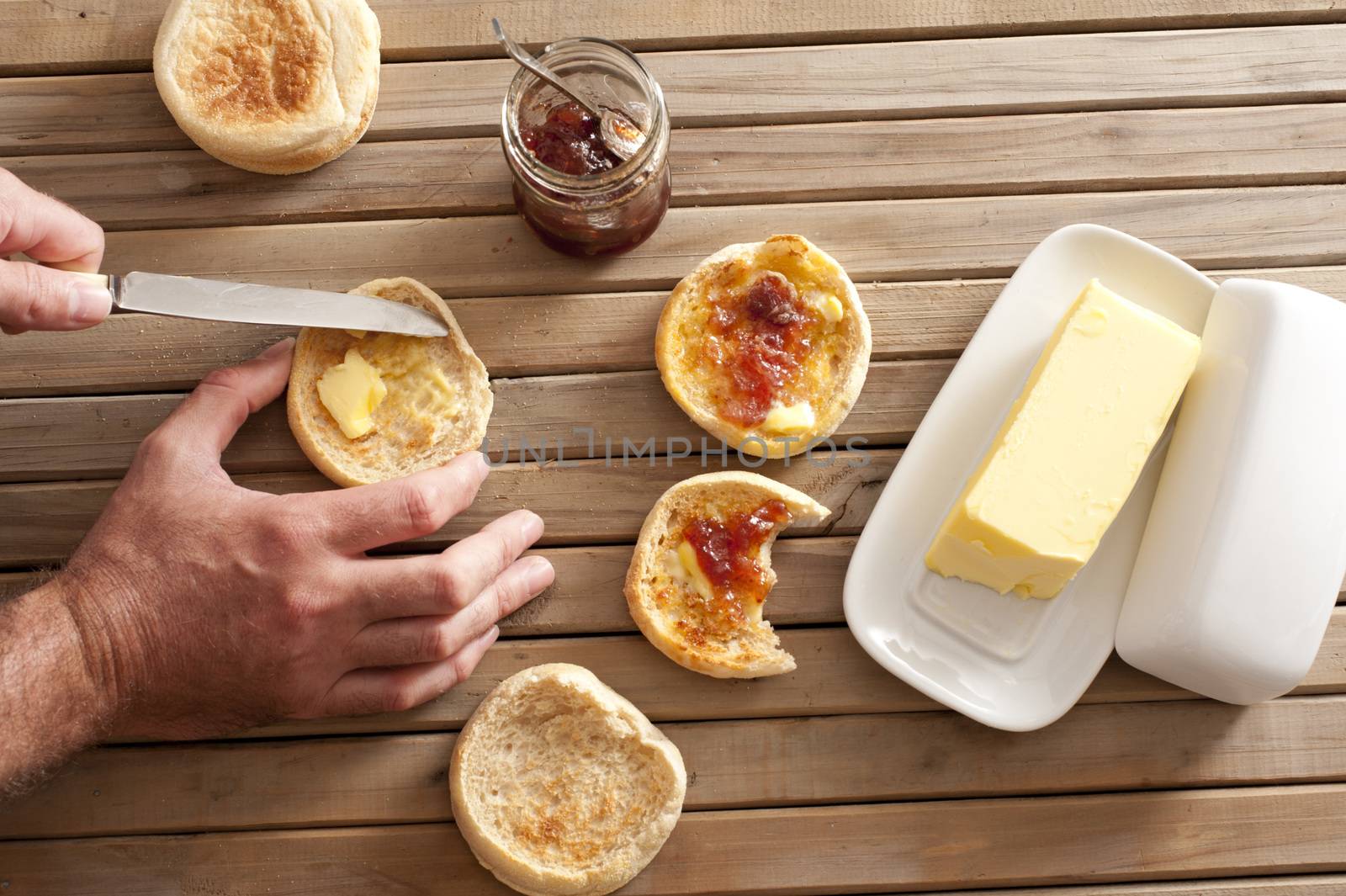 Man buttering a freshly toasted crumpet for breakfast, overhead view of his hands, the crumpets, jam and a pat of farm butter on a slatted wooden table