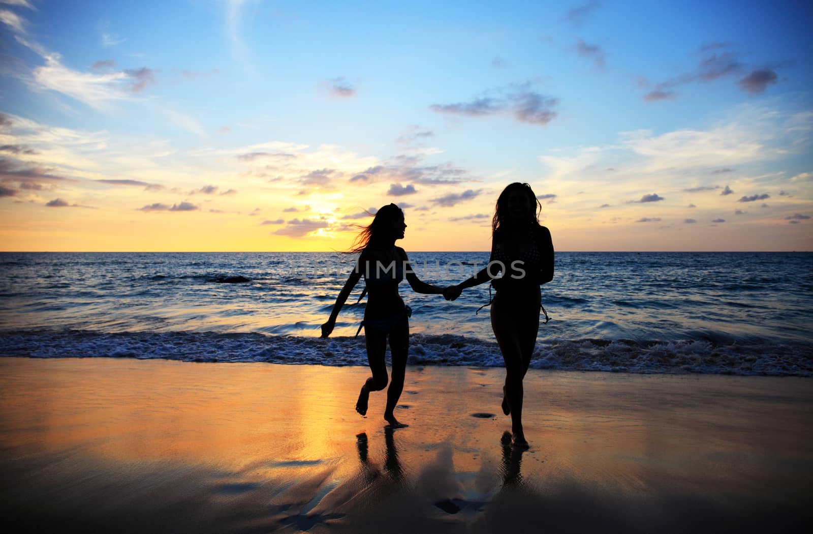 Two young happy women walking on tropical sea beach at sunset