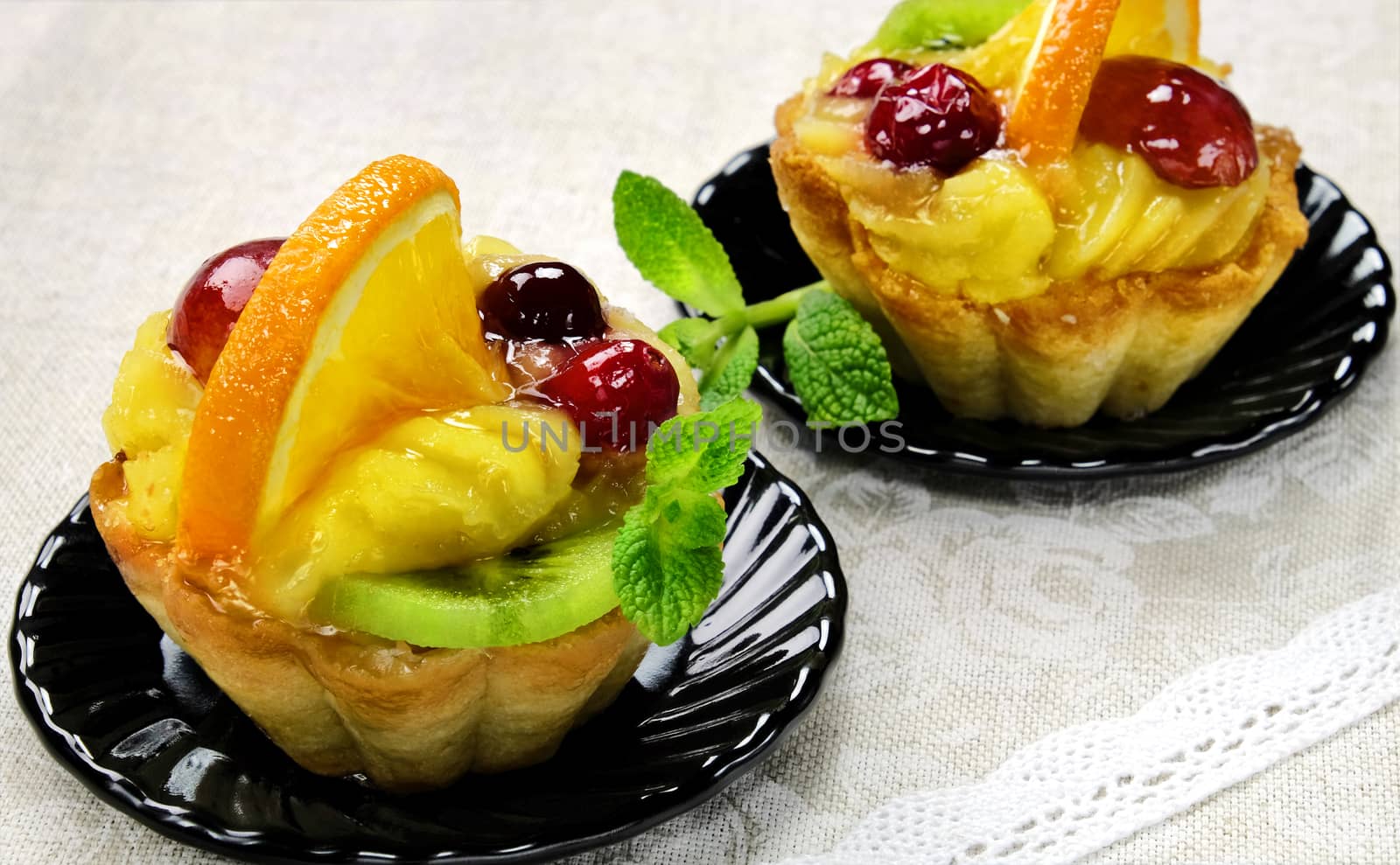Cakes with slices of fresh fruit orange, kiwi and grapes with jelly and cream on a black saucers on a table cloth.