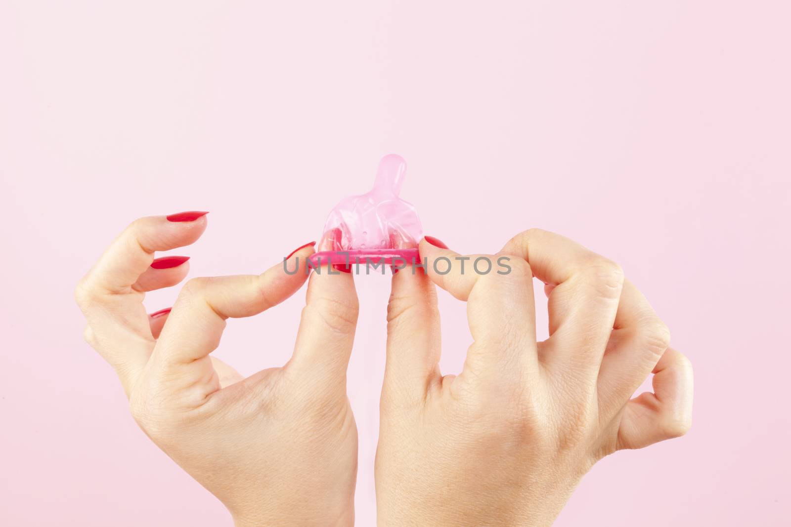 Female hands with red nails holding a pink condom isolated on pink background. Safe sex and birth control concept. 