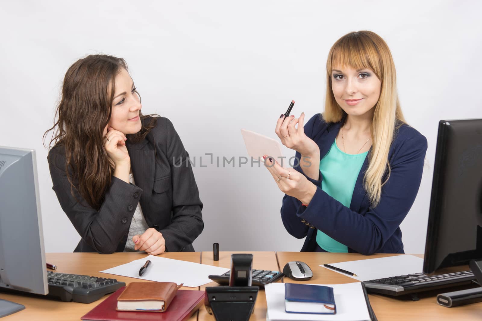 Office employee at his desk looking at his colleague, who is holding lipstick and mirror in hand