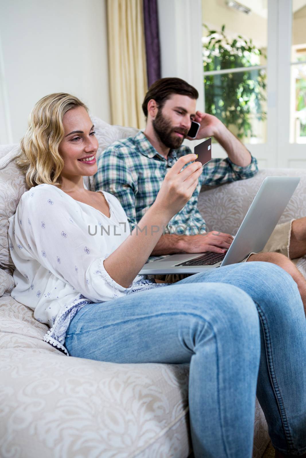 Woman holding credit card while sitting by man talking on phone with laptop at home