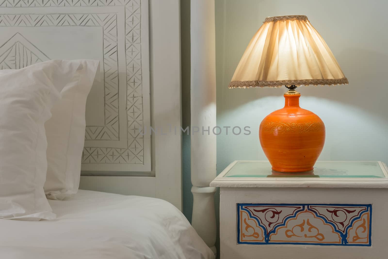 A lamp on a nightstand next to a bed with a white carved headboard