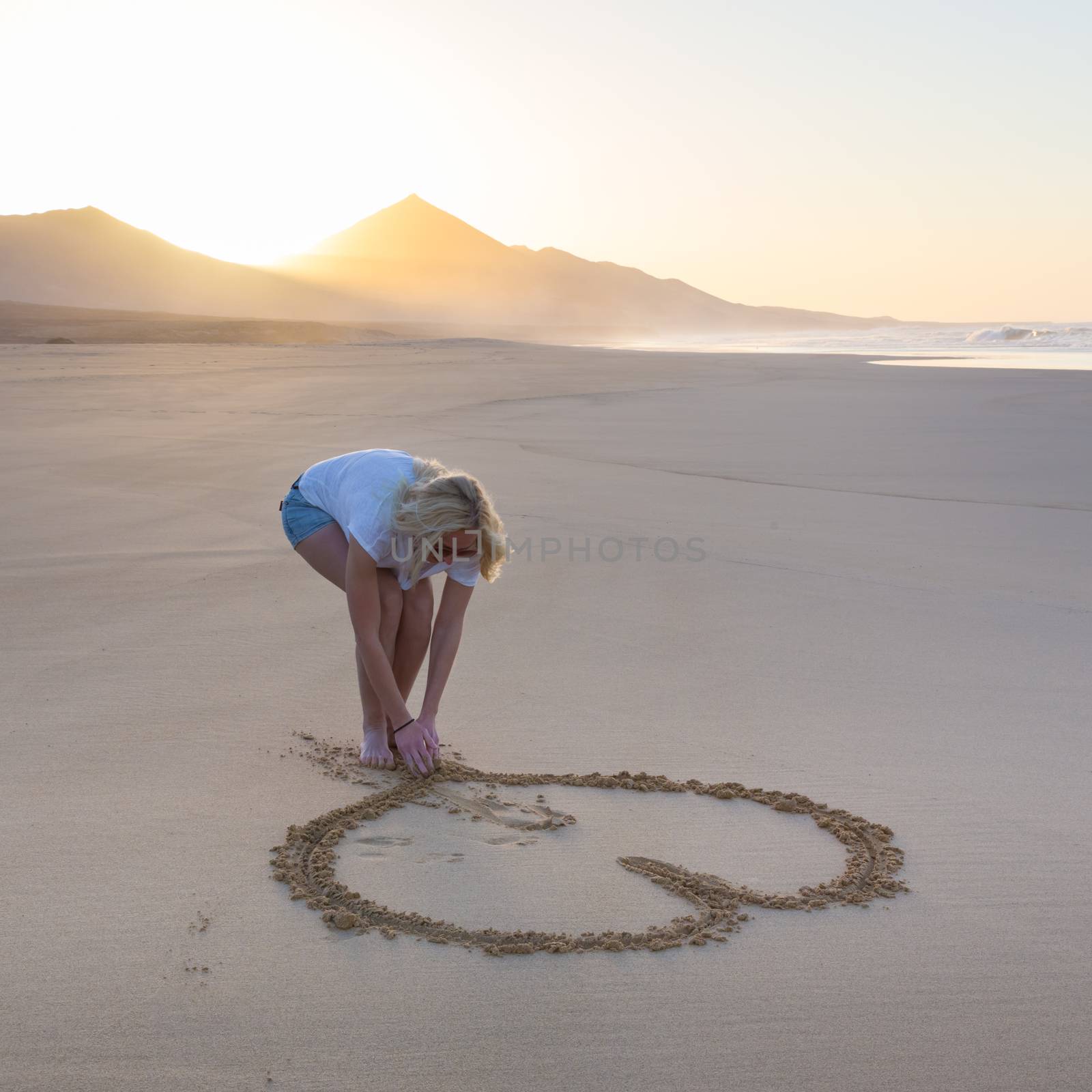 Woman drawing heart in sand on beautiful solitary Cofete beach in sunset. Love, travel concept. Fuerteventura, Canary Islands, Spain.